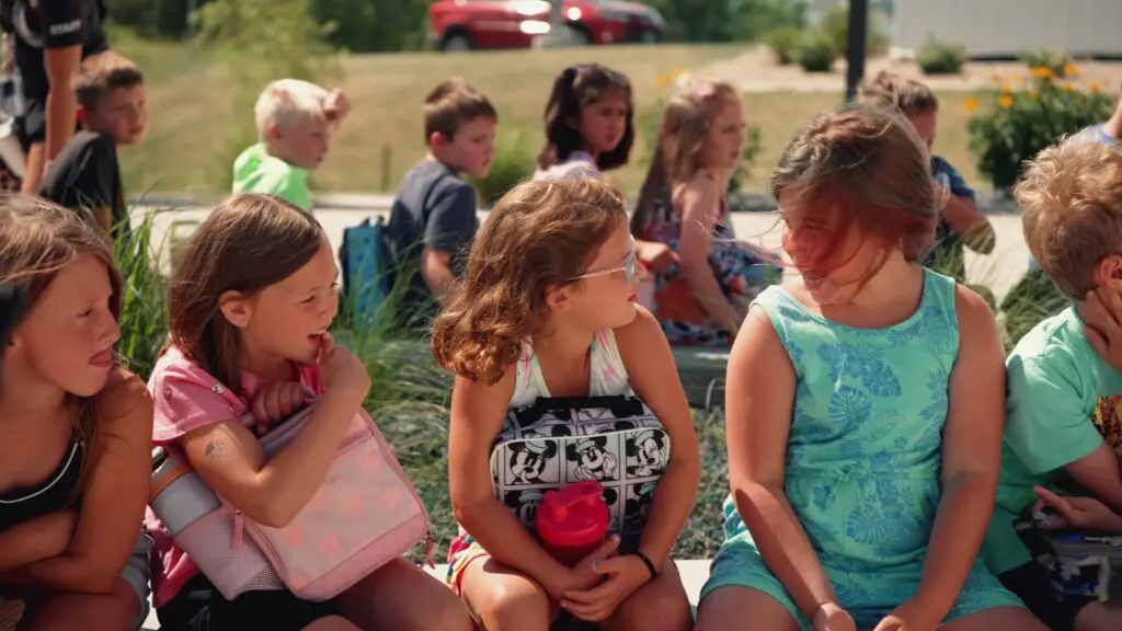 Children sitting outside in a row holding lunchboxes and talking together on a sunny day
