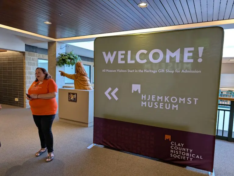 A woman in an orange shirt speaks beside a welcome sign at the Hjemkomst Museum