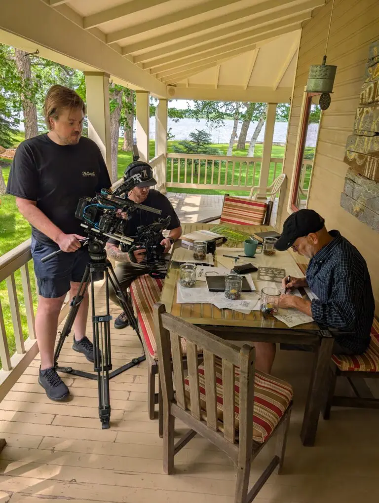 A film crew records a man writing at a table on a front porch