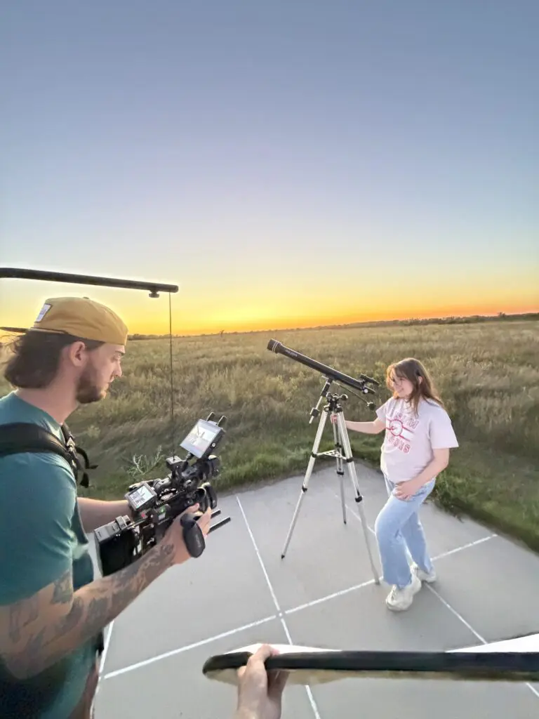 A videographer films a young woman beside a telescope at sunset