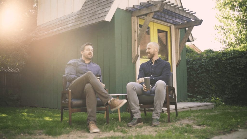 Two men sitting in chairs outside a small house, part of Craftwell Architecture's reintroduction to the community.