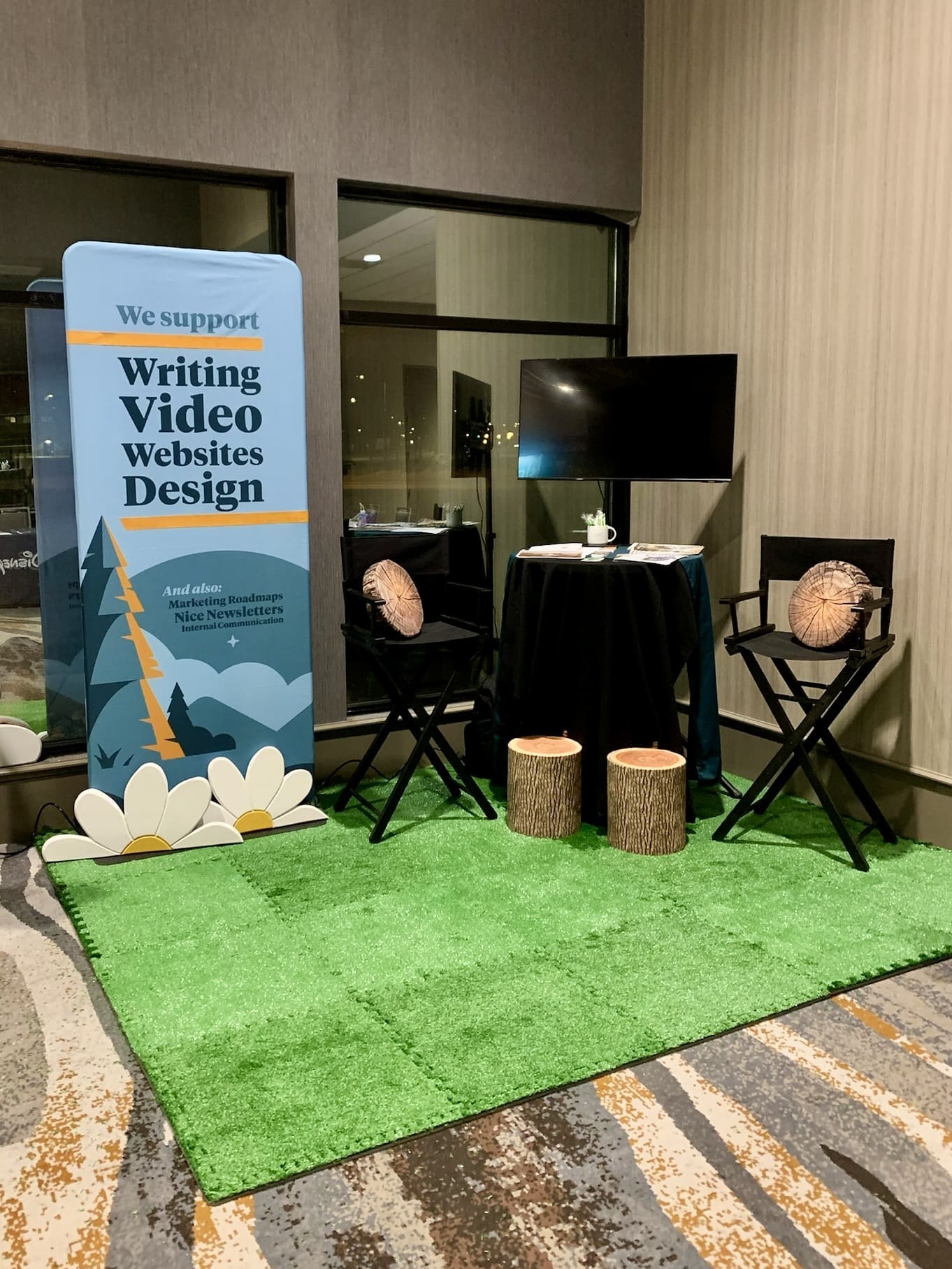 An exhibit booth at an indoor event featuring a vertical banner with text 'We support: Writing, Video, Websites, Design' and a list of other services. The booth has a faux grass area with two director's chairs and tree stump stools. A large flat-screen TV is turned off, mounted on a stand. The area is warmly lit, and reflections are visible in the glass window, indicating an evening setting.
