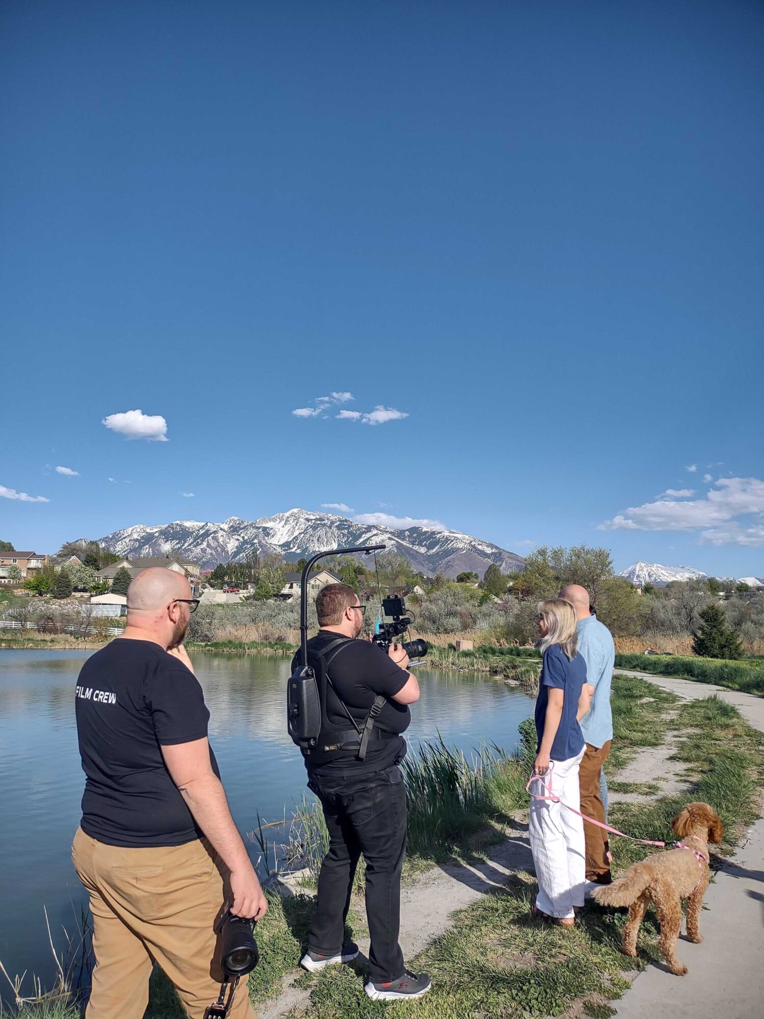 Outdoor shot of a film crew member filming a couple walking their dog by a pond, with the film crew's assistant looking on and mountains in the backdrop.
