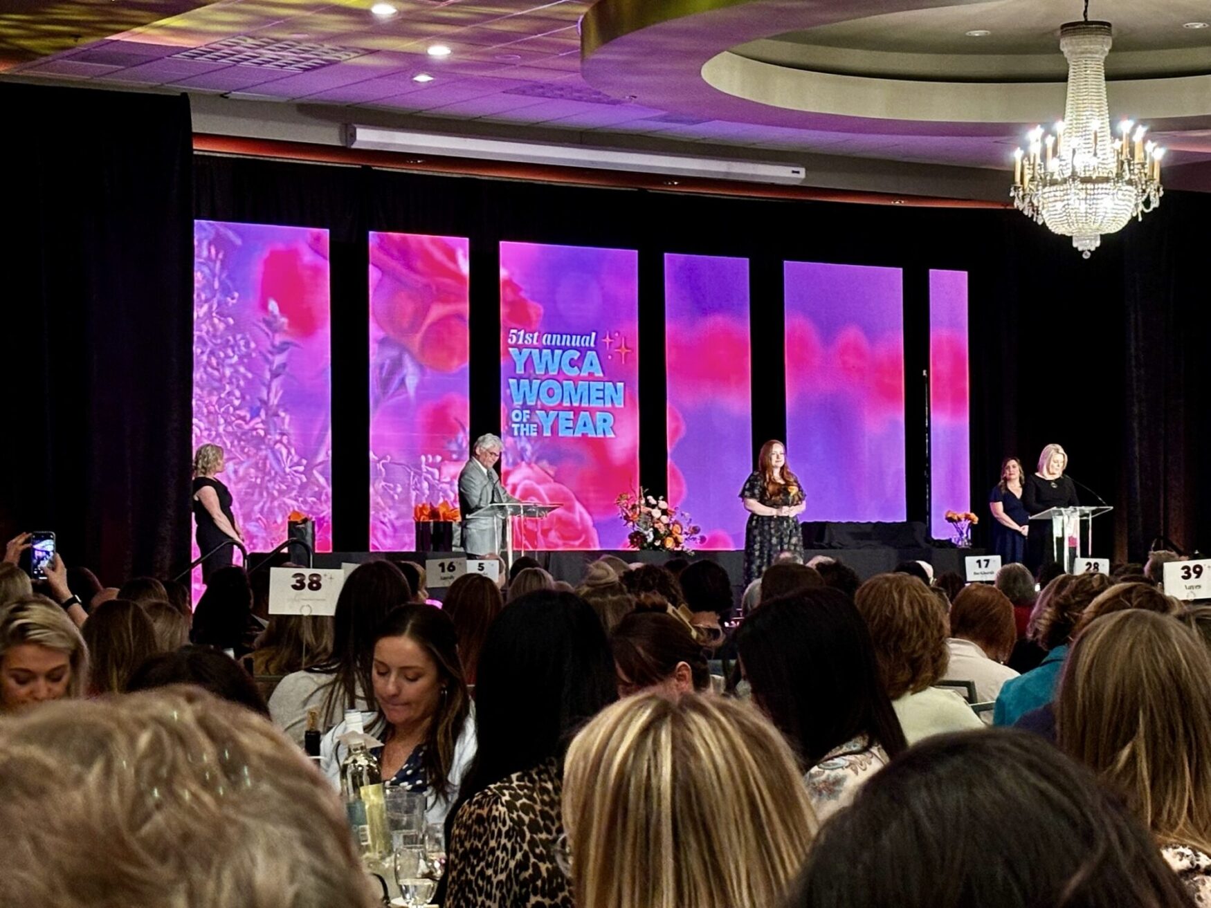 Indoor event scene showing a speaker at a podium during the 51st annual YWCA Women of the Year awards, with attendees in the foreground and colorful screens in the background.