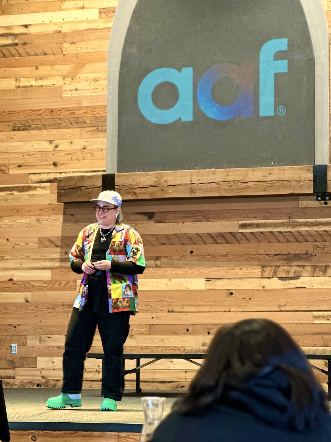 A speaker stands in front of a large American Advertising Federation (AAF) logo displayed on a wood-paneled wall. The speaker is wearing a colorful buttoned shirt, black trousers, and playful green monster feet slippers. They are smiling, seemingly engaged in a light-hearted moment. In the foreground, the back of an audience member's head is visible, suggesting an informal and interactive event atmosphere.