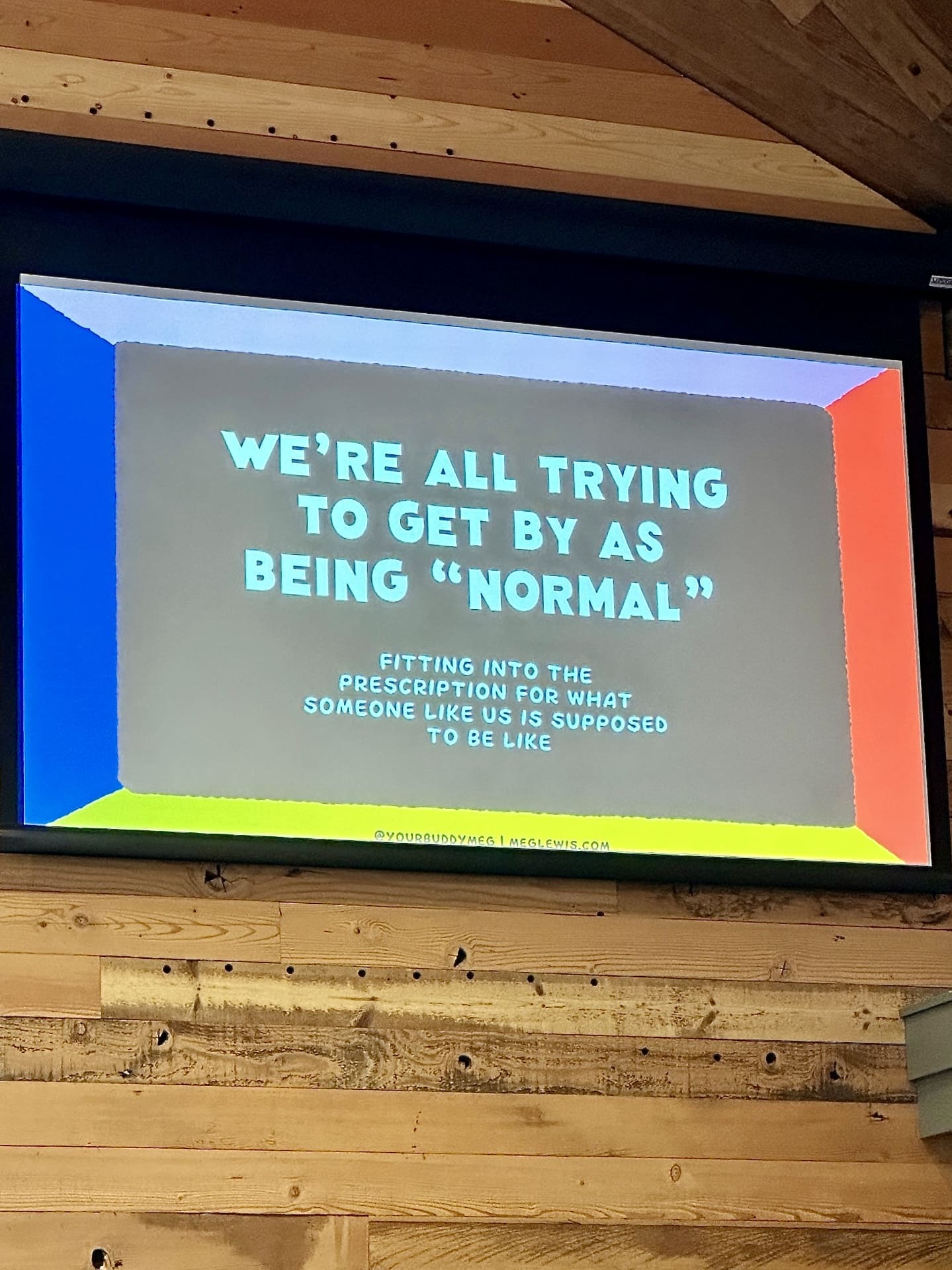 A large, colorful presentation slide projected onto a screen with a wooden beam ceiling above. The slide's background is segmented into bright blue, green, and red sections, with the following text in bold, shadowed letters: 'WE'RE ALL TRYING TO GET BY AS BEING "NORMAL" FITTING INTO THE PRESCRIPTION FOR WHAT SOMEONE LIKE US IS SUPPOSED TO BE LIKE.' Below, in smaller print, are the social media handle '@yourbuddymeg' and the website 'meglewis.com'.