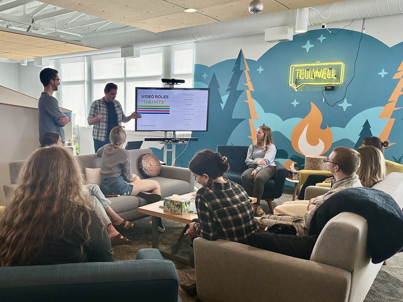 A casual office presentation with two speakers in front of a screen displaying 'VIDEO ROLES' while a group listens attentively, seated on couches in a cozy set up.