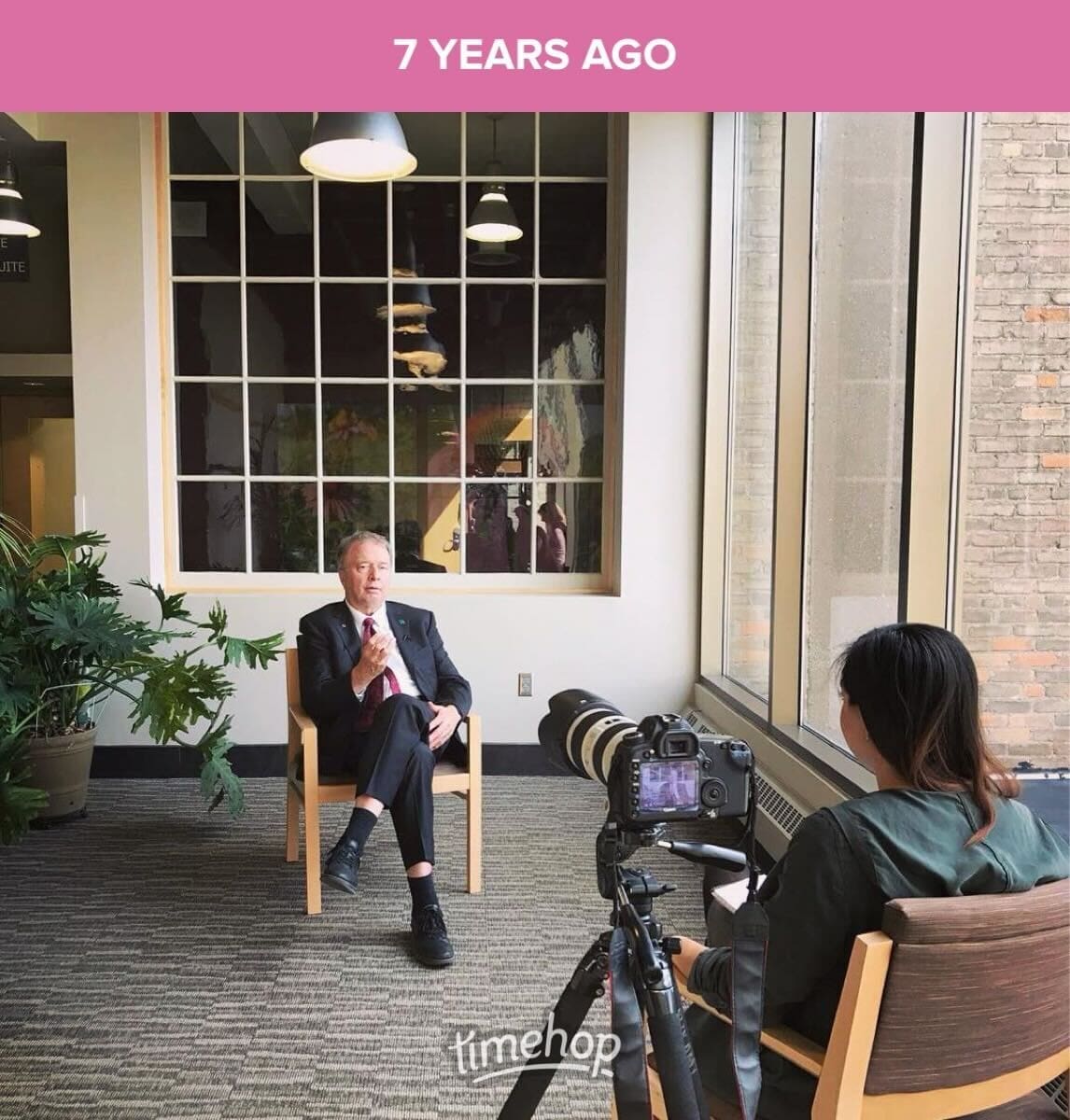 A Timehop photo showing a candid moment from 7 years ago. A man in a suit sits on a wooden chair in an indoor setting, gesturing mid-conversation. In front of him, a camera on a tripod is focused on him, operated by a person whose back is to the camera. The room is well-lit with natural light streaming in through a large window with white frames. A couple of potted plants add a touch of greenery to the scene.