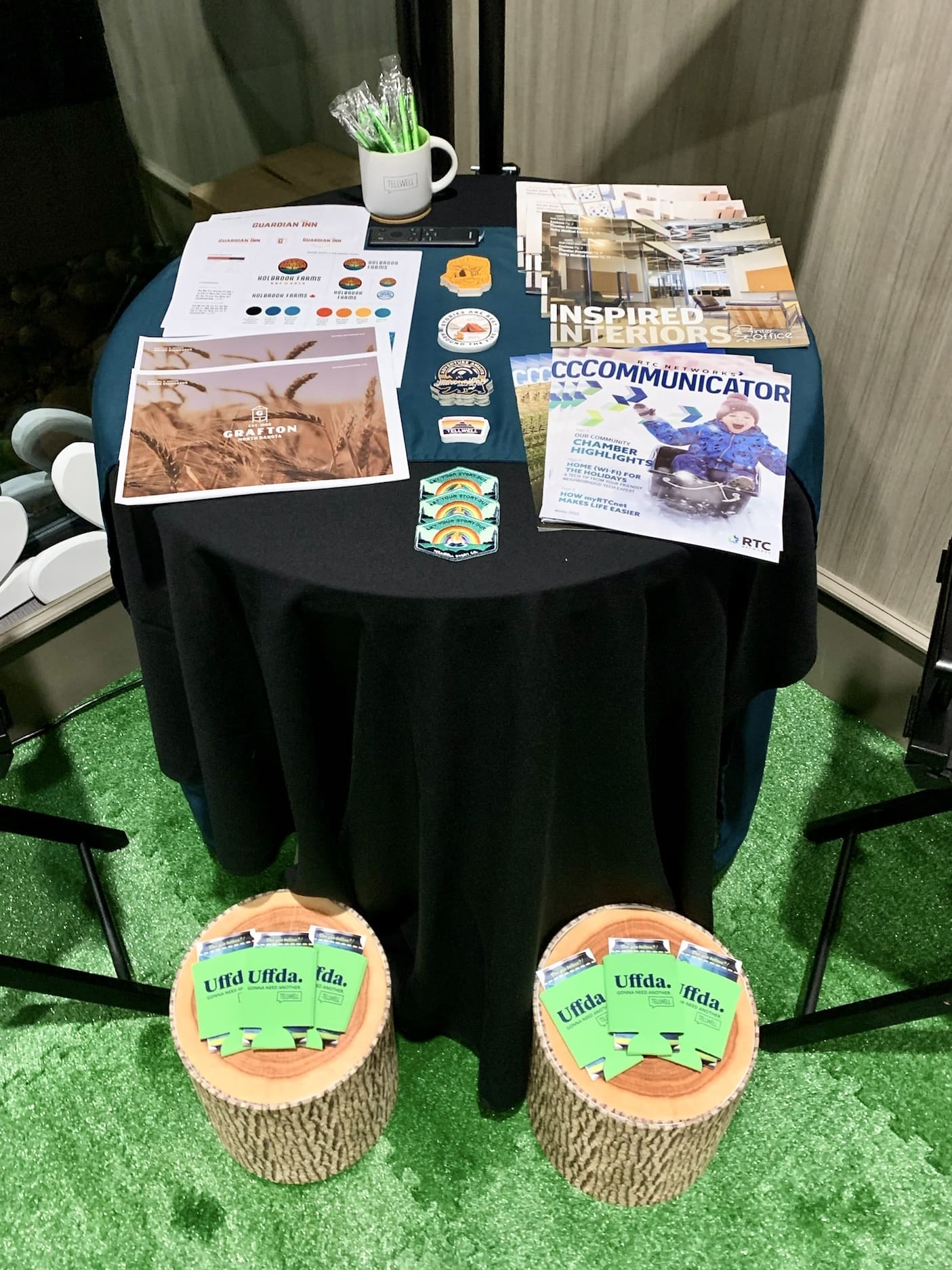 An informational table at an event with a black cloth covering, displaying various printed materials. There are brochures, pamphlets titled 'INSPIRED INTERIORS' and 'COMMUNICATOR', as well as a mug with pens and branded stickers. Two tree stump stools sit in front of the table, each with a stack of green 'Uffda' koozies on top. The setup is on faux grass flooring, suggesting an inviting, creative presentation.
