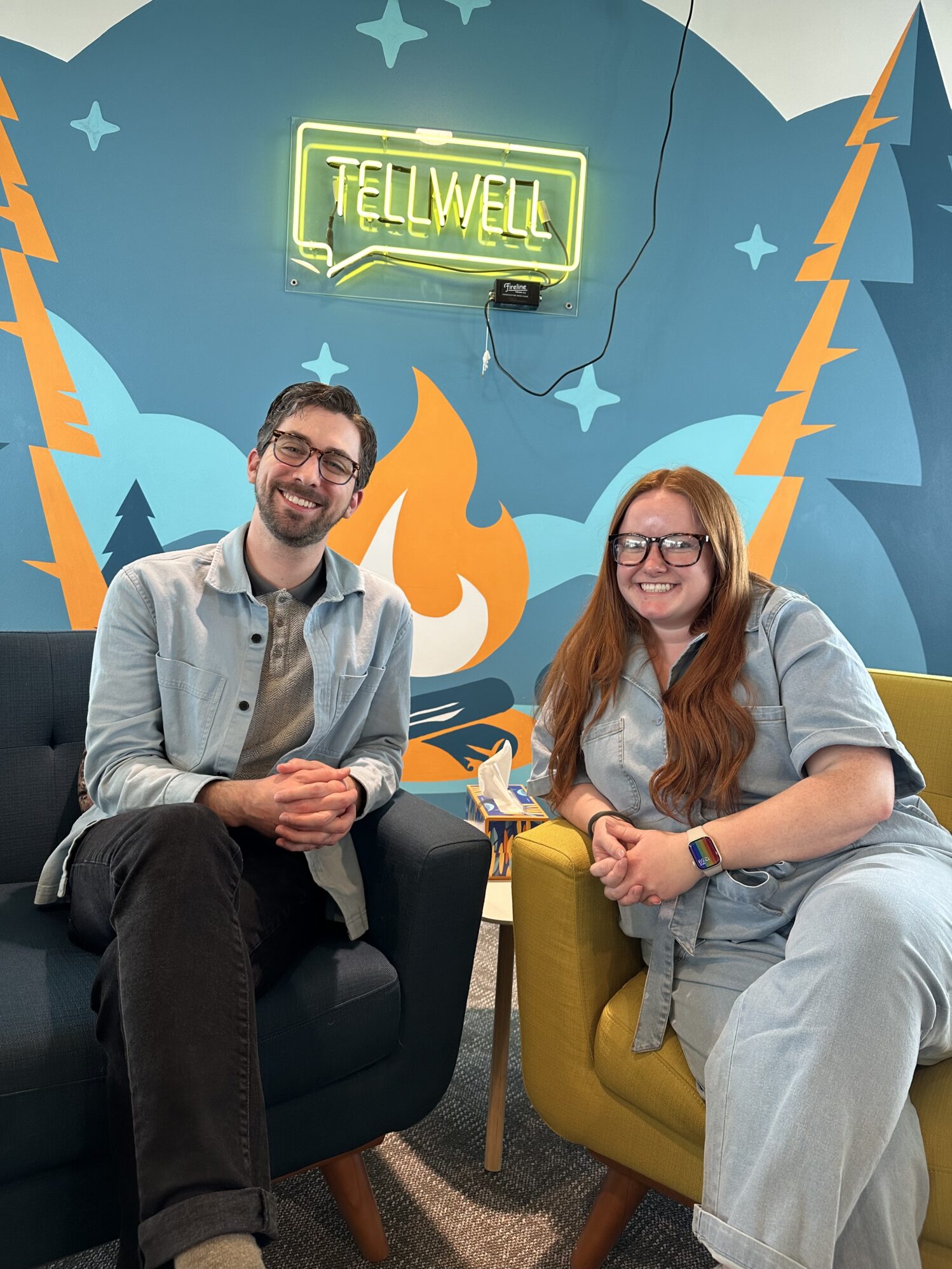 Two smiling individuals–wearing incredibly similar outfits– seated on a couch in a vibrant office environment with a neon 'TELLWELL' sign on the wall, portraying a welcoming and creative workspace.