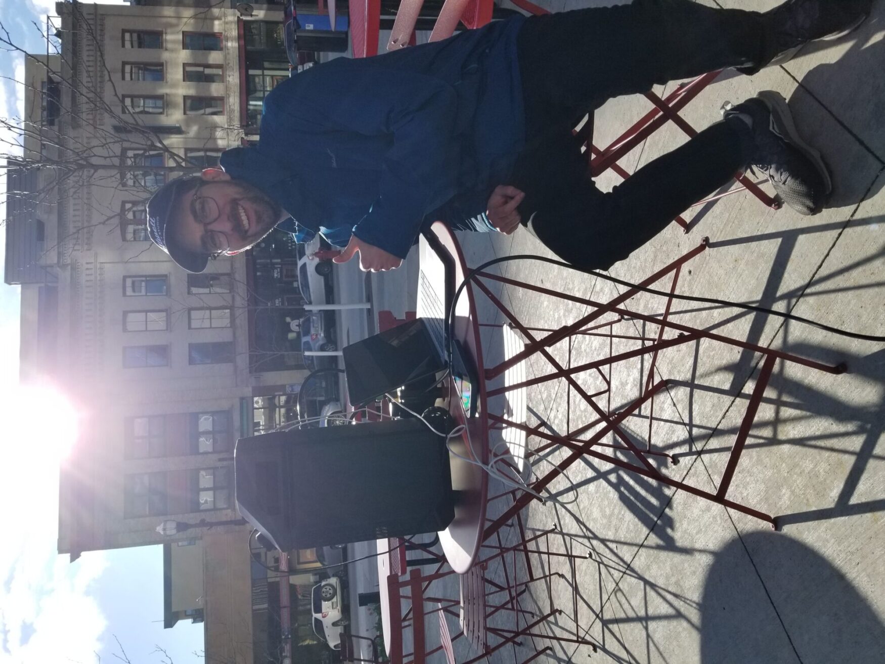 A man sitting at an outdoor cafe table giving a thumbs up. He's wearing a blue jacket and glasses, with a joyful expression, sitting in sunlight with urban buildings in the background.
