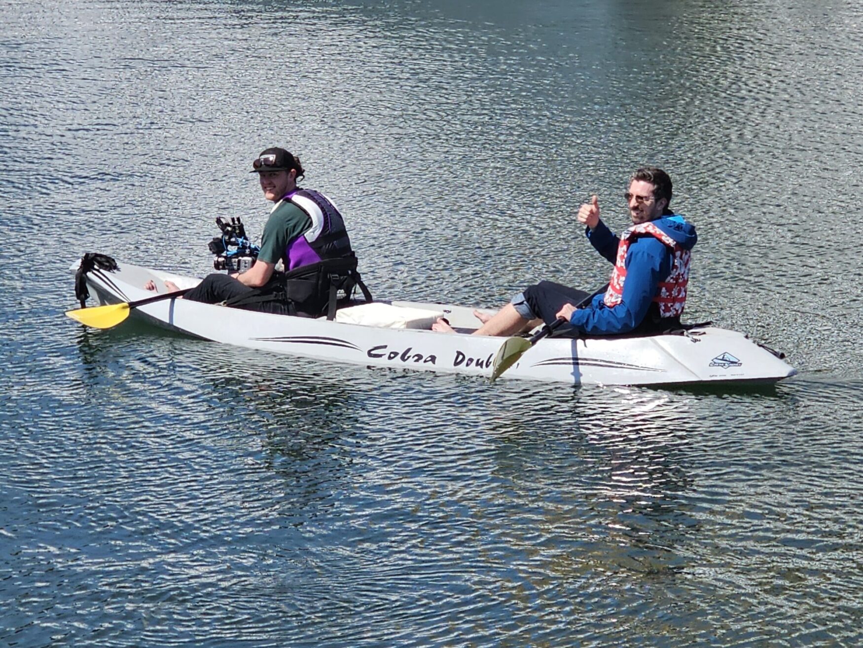 Two people are kayaking on a calm body of water. One person is holding a camera, while the other gives a thumbs-up. Both are wearing life jackets.