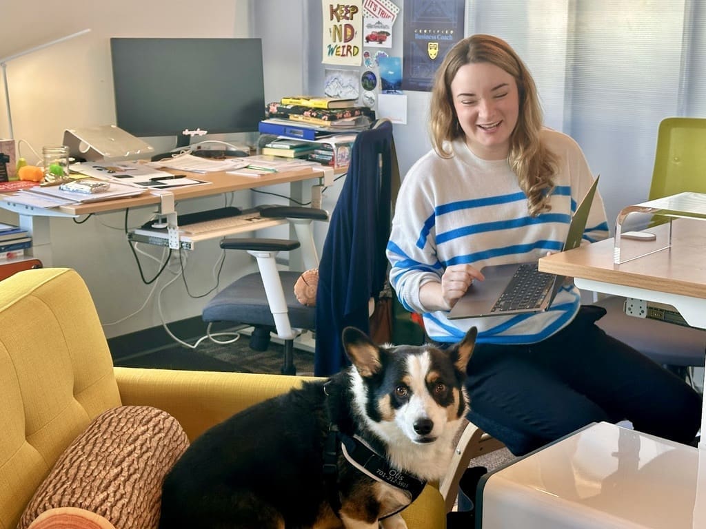 A woman with long blonde hair, wearing a white and blue striped sweater, sits at a desk holding a laptop and smiling. She is in a cozy, cluttered office space with a variety of items on the desks and shelves, including books, a lamp, and a computer monitor. Next to her, sitting on a yellow armchair, is a black and white Corgi dog with a harness. The atmosphere appears relaxed and friendly, suggesting a pet-friendly workplace.