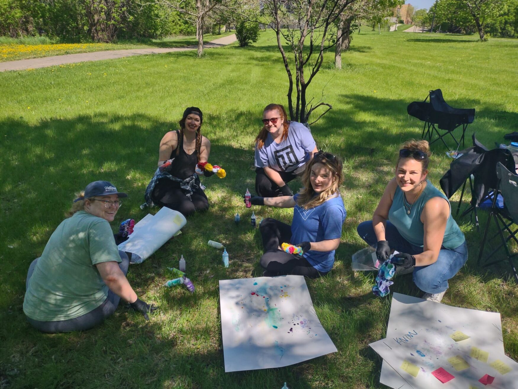 Five people are sitting on the grass in a park, engaged in a tie-dye activity. They are smiling and holding bottles of dye, with tie-dyed items and materials spread out on the ground. Trees and a walking path are visible in the background.