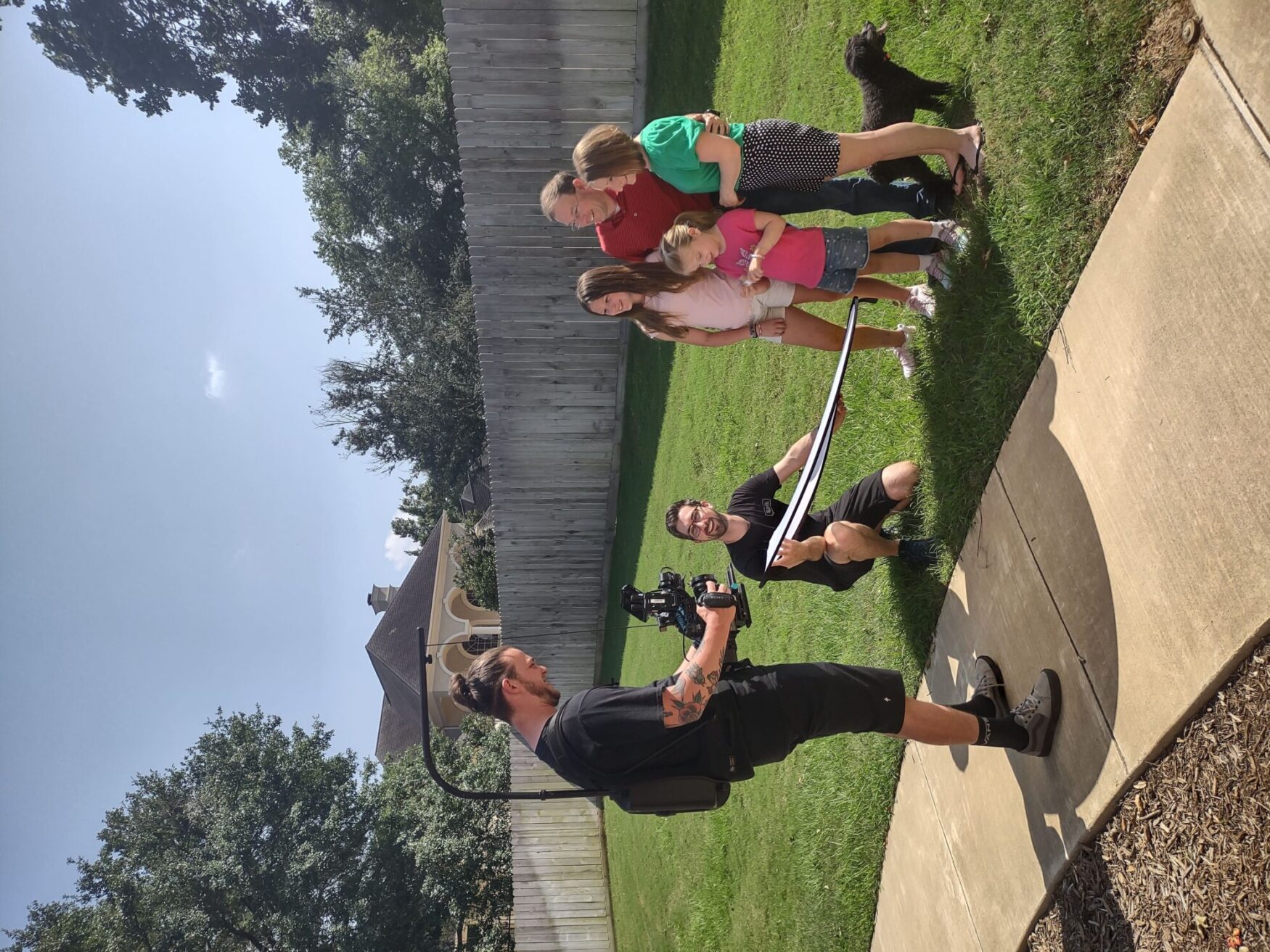 A man is filming a family of four, who are standing in a yard. The family includes two adults and two children, along with a dog. The cameraman and another person are setting up the shot.