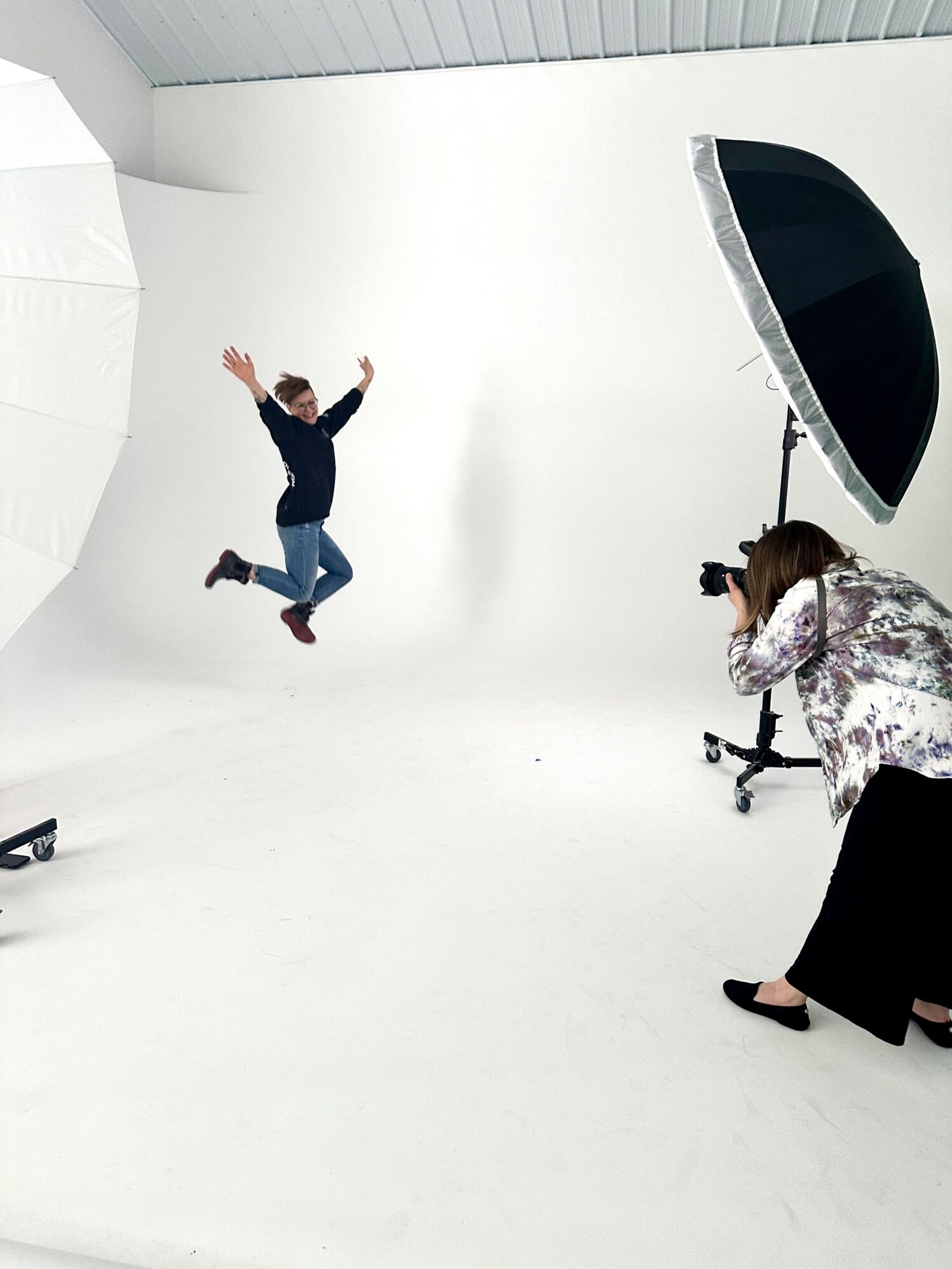 A woman jumping high with joy during a photoshoot in a studio setting, surrounded by white walls and professional lighting equipment. The photographer is also in the photo, using a large camera to capture the subject’s jump.