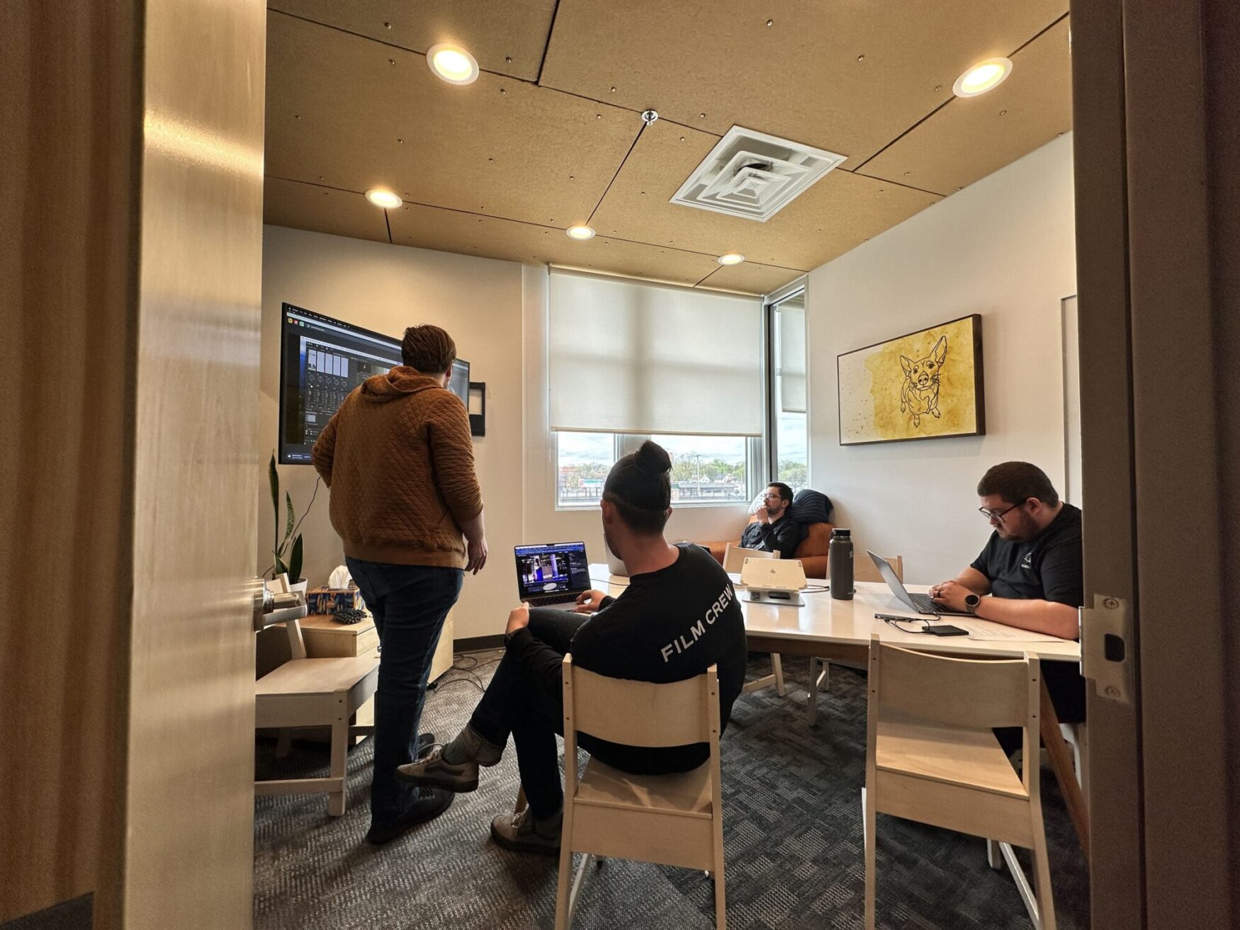 An indoor setting with a film crew at work. One person is looking at a computer screen showing video editing software, and others are focused on their laptops, in a well-lit room with modern decor.