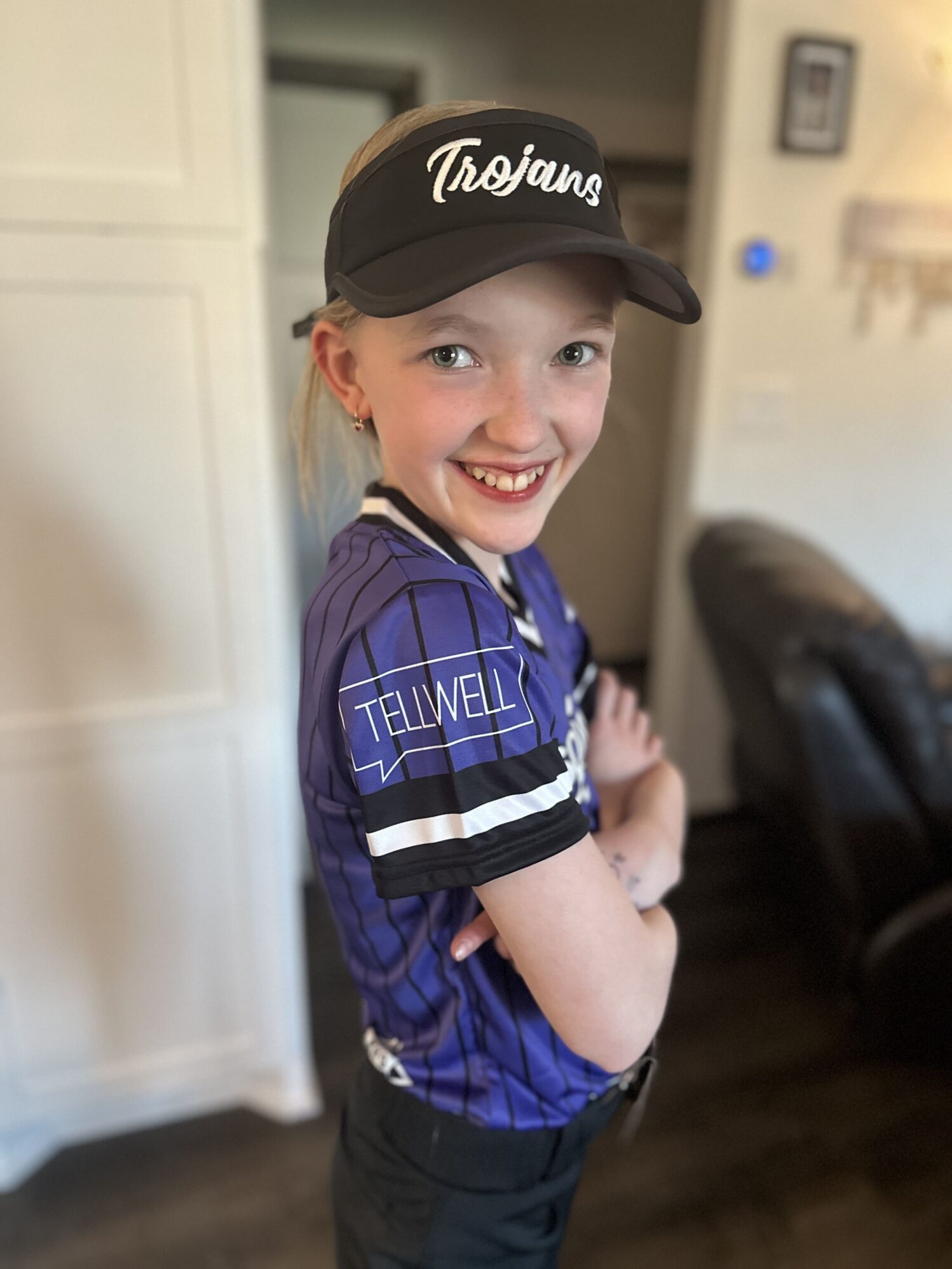 A young girl wearing a sports uniform and a baseball cap. She has a cheerful smile and is posing with her left hand on her hip. The cap is labeled "Trojans" in cursive script, and her jersey is purple with white and black accents and the name "TELLWELL" across the chest. Her expression and posture suggest confidence and happiness.