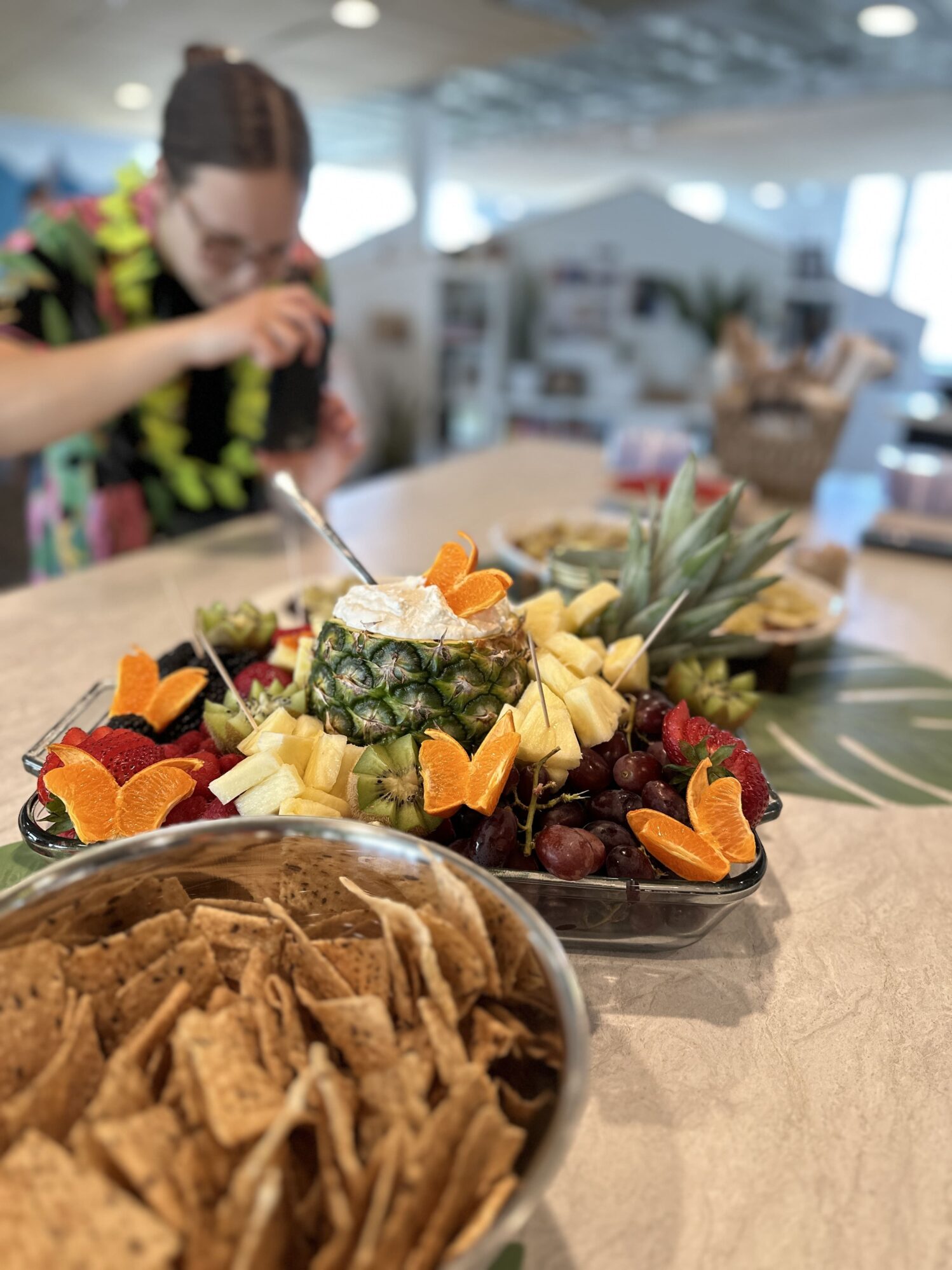 A food table with a tropical theme, featuring a pineapple, mixed fruits, and chips. A young woman in a bright floral shirt is serving food, slightly blurred in the foreground, emphasizing the colorful and arranged fruits.