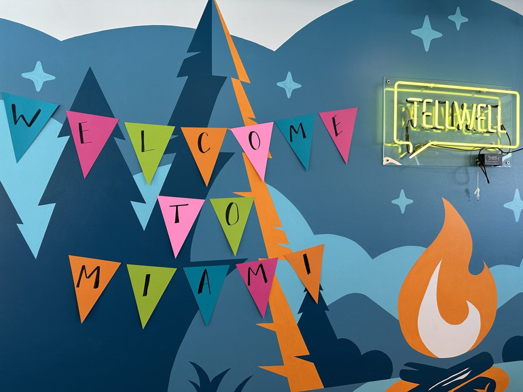 A vibrant wall mural featuring stylized mountain peaks and waves with a "Welcome to Miami" message in colorful triangular banners. A neon sign that reads "Tellwell" is mounted on the right side of the mural.