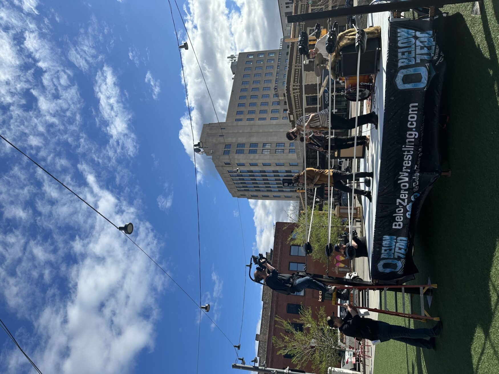 A band performs on a wrestling ring repurposed as a stage in an urban outdoor setting. A videographer on a ladder captures the performance, with city buildings and a clear sky in the background.