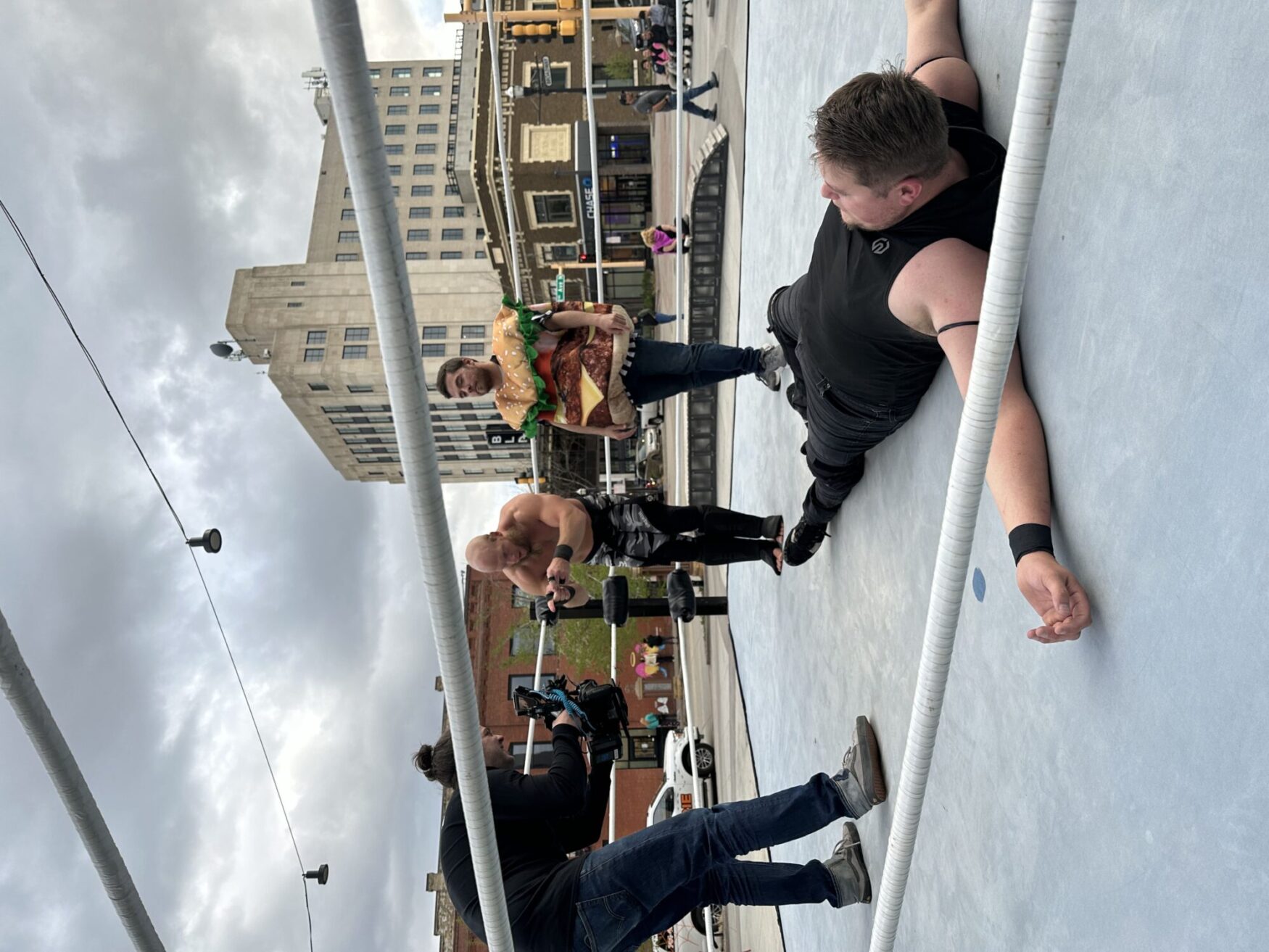 Two wrestlers in a ring, one dressed as a hamburger, during an entertaining match, while a cameraman captures the scene, all set against an urban backdrop under a cloudy sky.