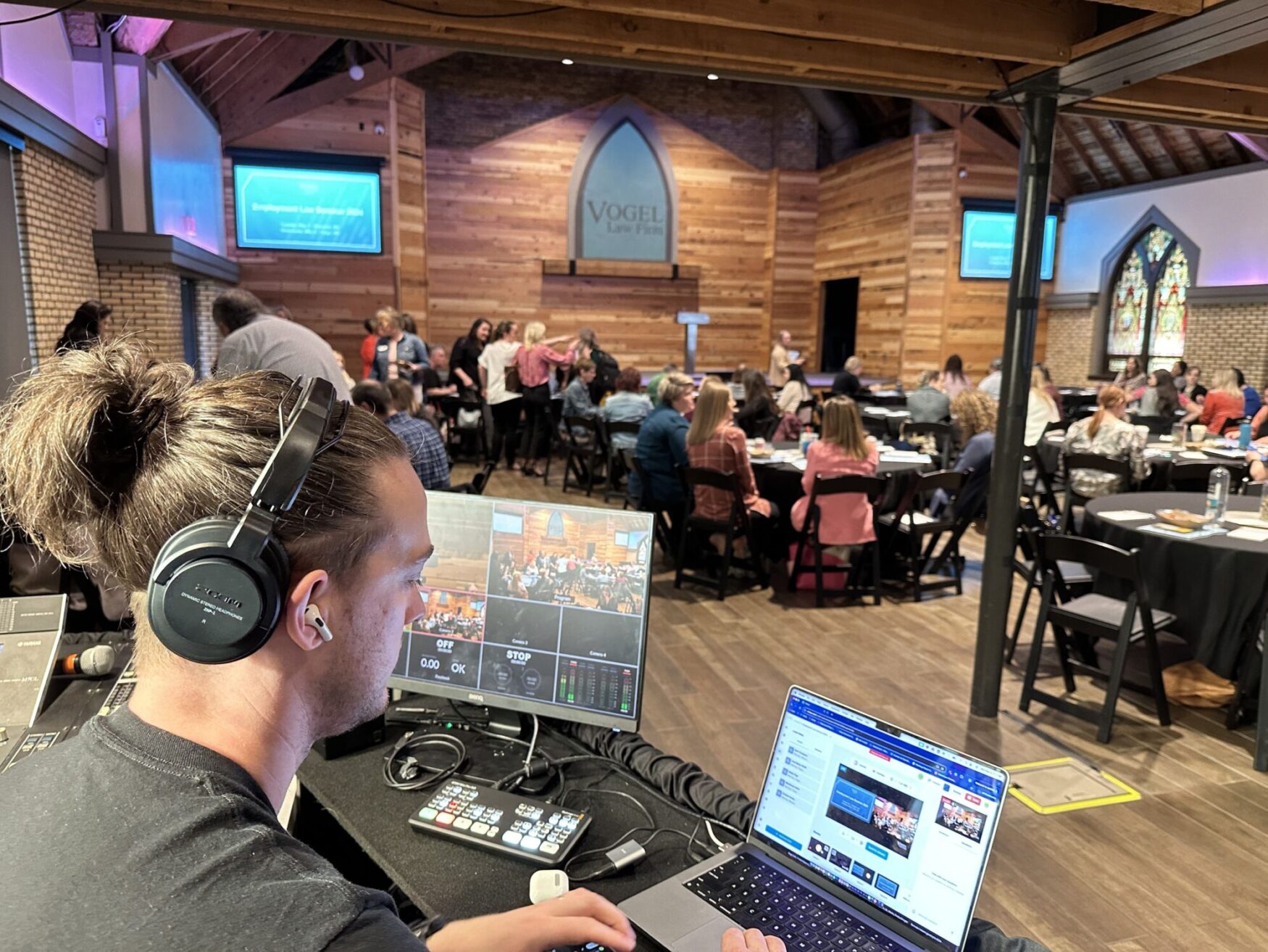 This image shows a man seated at a control desk, managing technical equipment during a live event. He is wearing headphones and focused on a laptop screen displaying multiple video feeds of the event. In front of him are various pieces of audio and video control gear. The background features a group of people seated at tables in a venue that combines modern elements with traditional architectural features, such as wooden beams and a stained glass window. The screen behind the audience displays "Employment Law Seminar 2020," indicating the nature of the event.