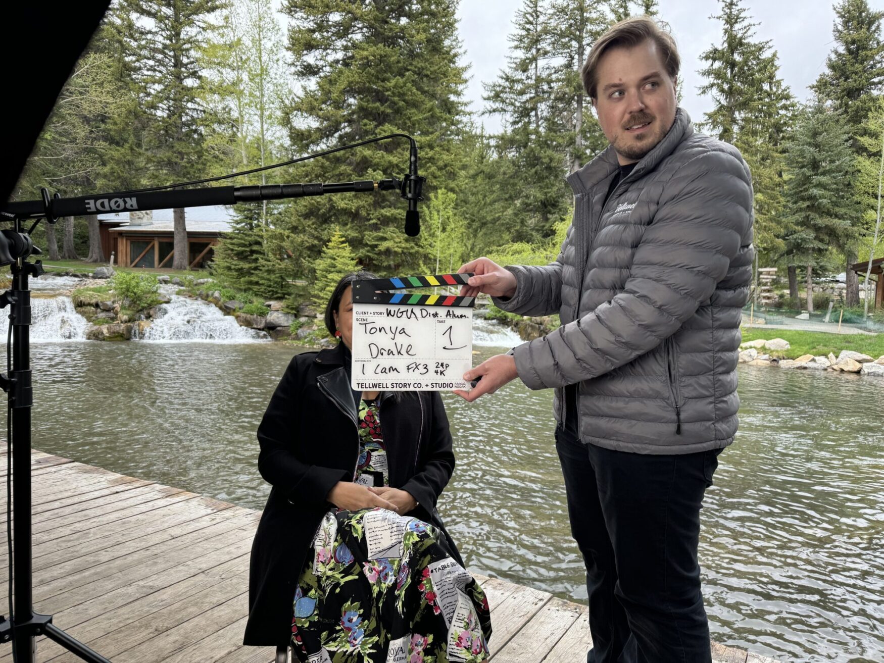 A man holds a clapperboard in front of a seated woman on a wooden deck beside a stream. The woman is partially hidden by the clapperboard, and a waterfall is visible in the background.