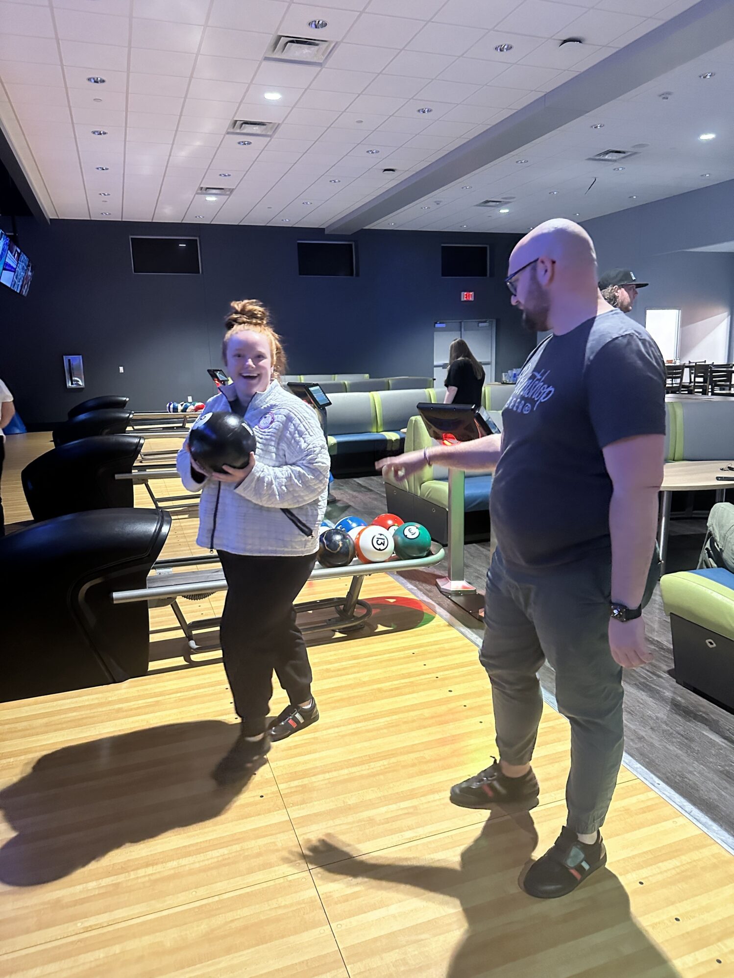A woman holds a bowling ball, smiling and looking towards a man who is instructing her. They are in an indoor bowling alley with other people visible in the background.