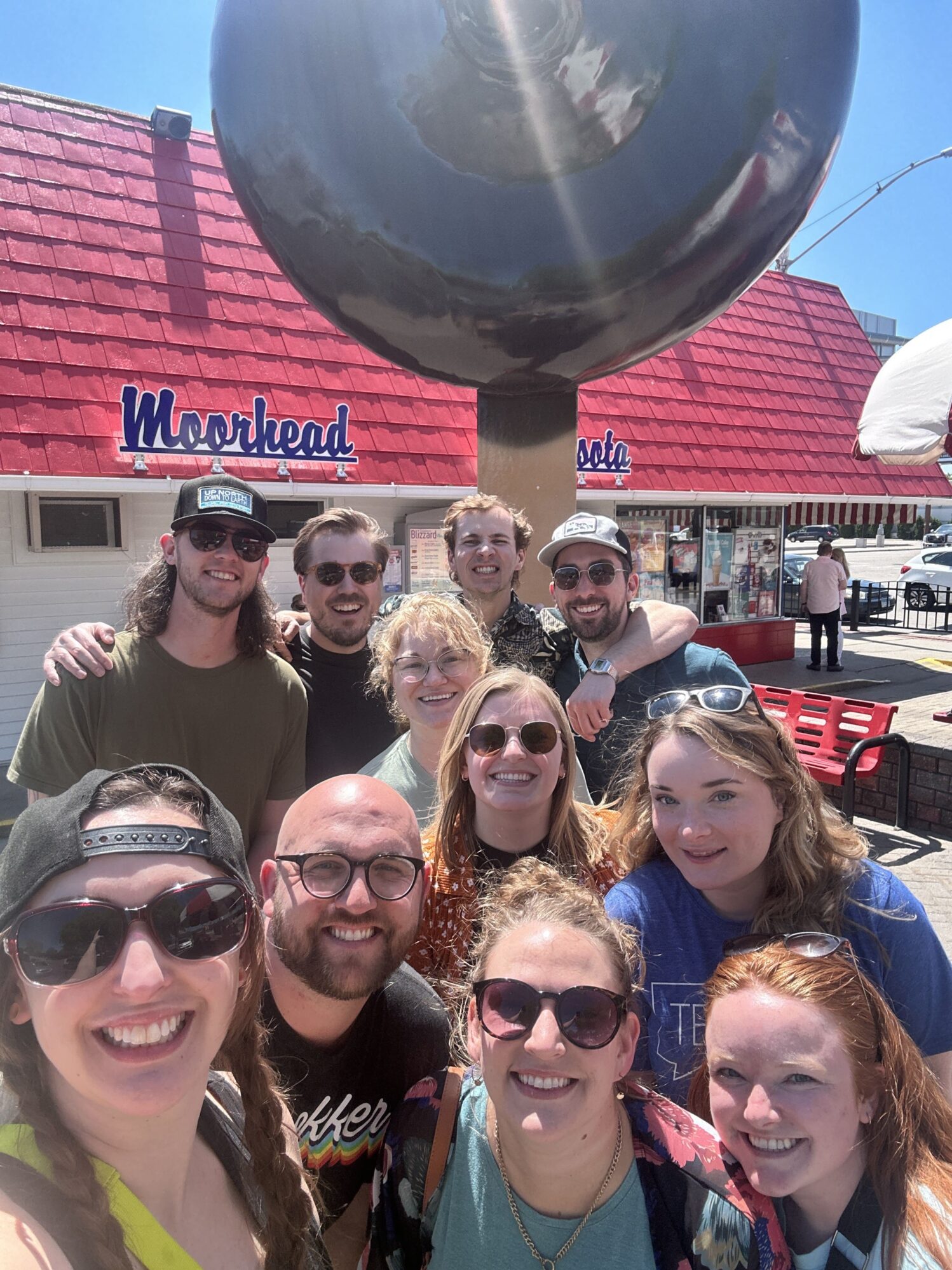 A group of nine people is taking a group photo in front of a large Dilly Bar sculpture. They are all smiling and enjoying a sunny day.