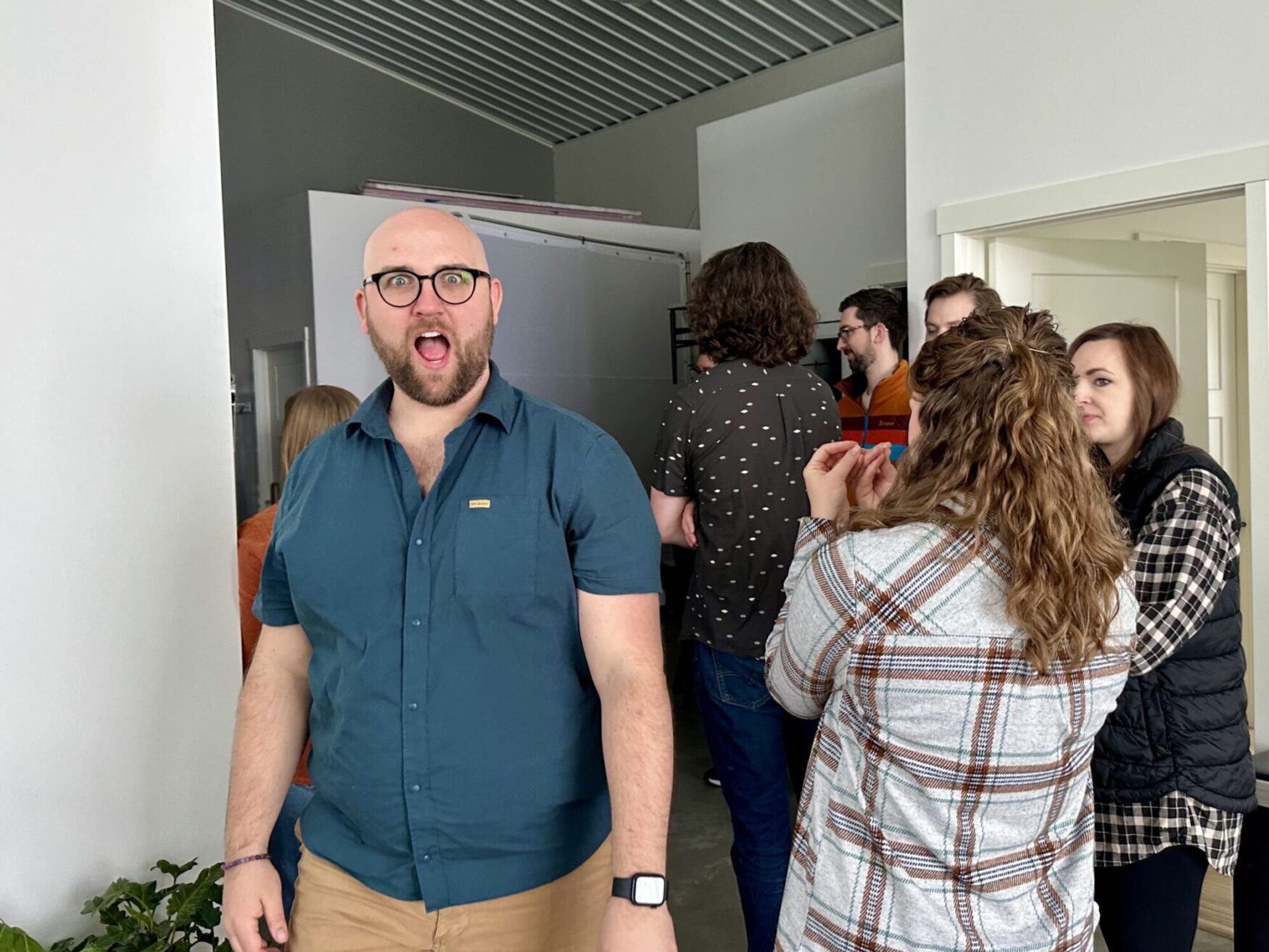 A man making a playful face at the camera in a photo studio with other people engaging in conversations in the background.