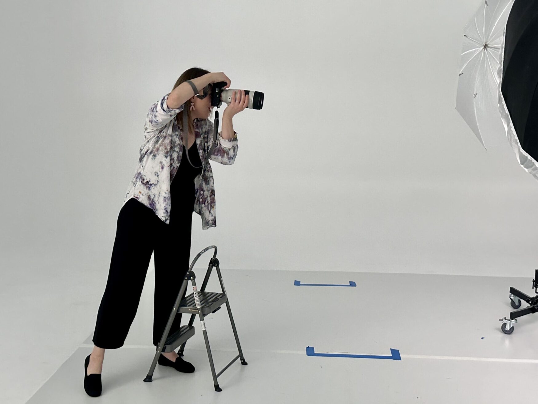 A female photographer focusing intently on taking a photo, with a large umbrella light stand beside her in a professional photo studio.