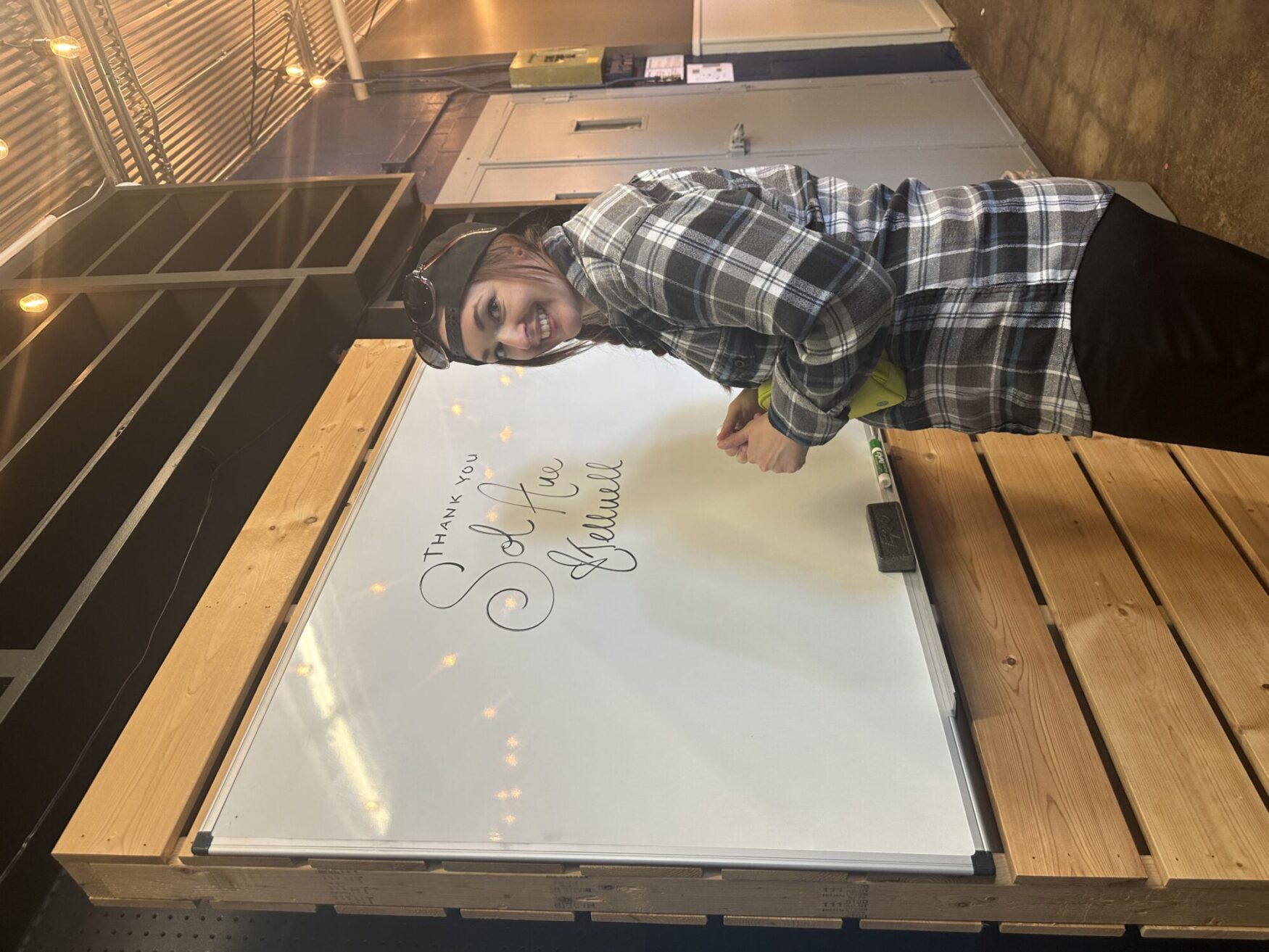 A woman wearing a plaid shirt and a backwards cap is writing on a whiteboard. The text on the whiteboard says, "Thank you Sol Ave and Tellwell." The setting appears to be indoors with string lights and a warm, rustic atmosphere.