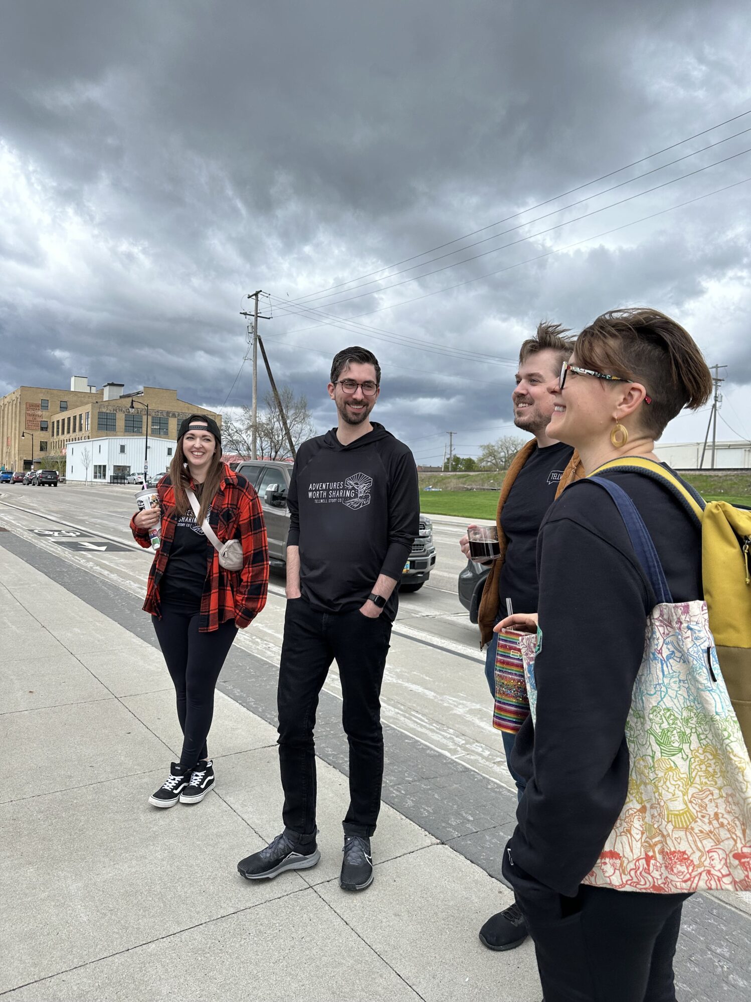 Four friends standing and chatting on a city street, smiling and dressed casually, under a cloudy sky, conveying a relaxed, friendly atmosphere.