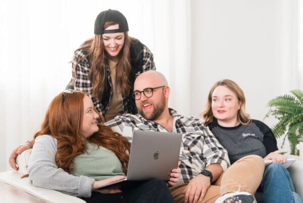 Four people are sitting and interacting on a white couch. A man with a beard and glasses holds a laptop, smiling and talking to a red-haired woman next to him. Another woman with long hair and a backwards baseball cap is standing behind them, smiling and looking down at the group. A fourth person with shoulder-length blonde hair is seated at the end of the couch, holding a notebook. A potted plant is visible in the background.