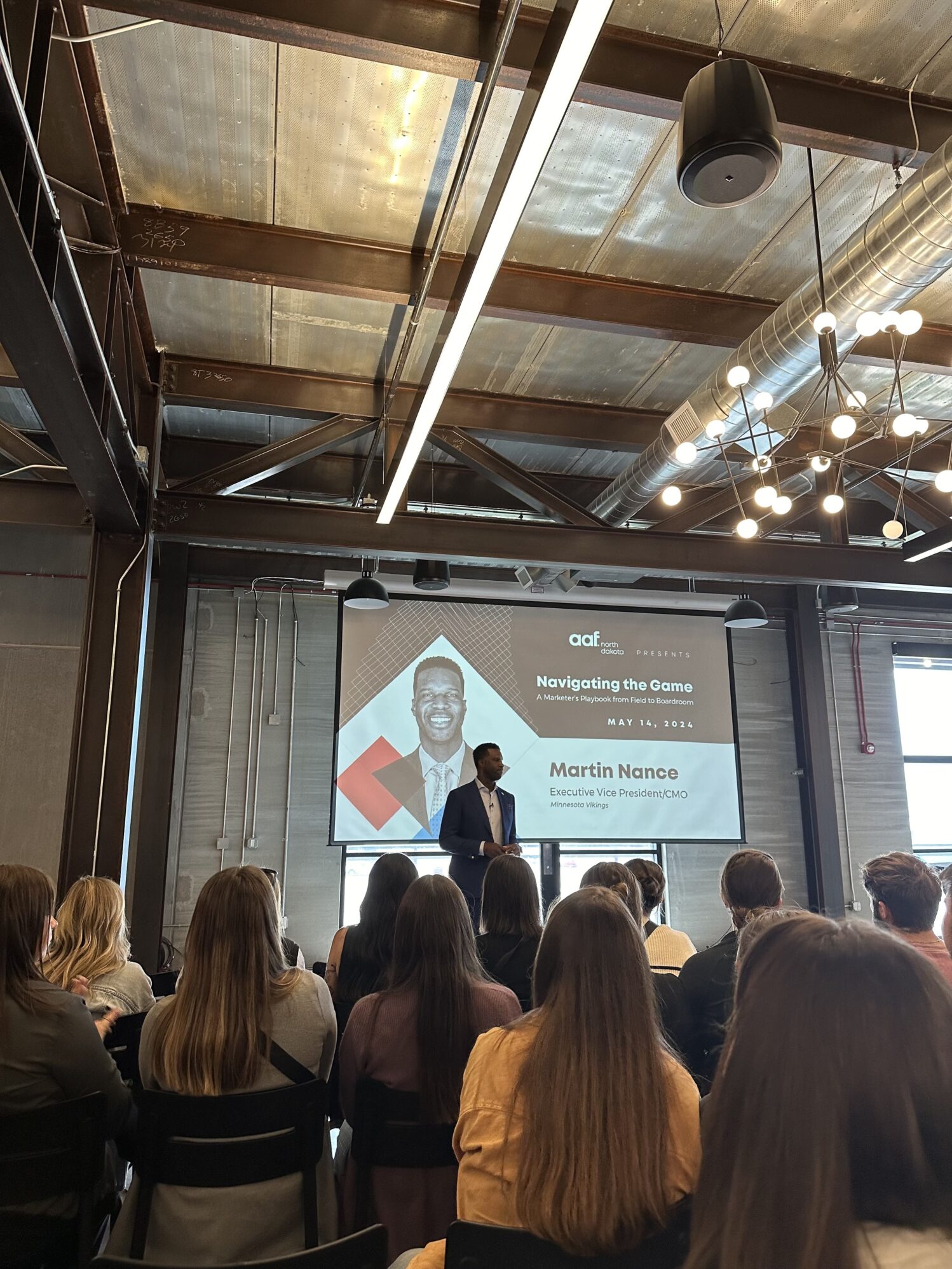 A man stands on a stage giving a presentation to an audience. The screen behind him displays his name, Martin Nance, and the title of the presentation, "Navigating the Game: A Marketer's Playbook from Field to Boardroom." The audience is seated and facing the stage in a room with industrial-style decor.