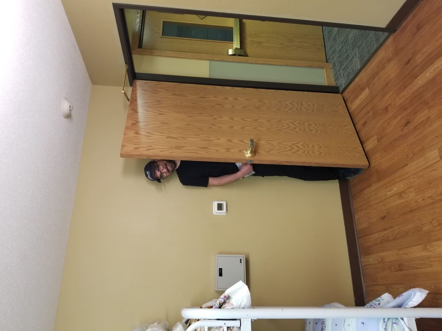 A man in a black shirt and cap stands partially behind a door, smiling in a room with bunk beds and wooden floors.