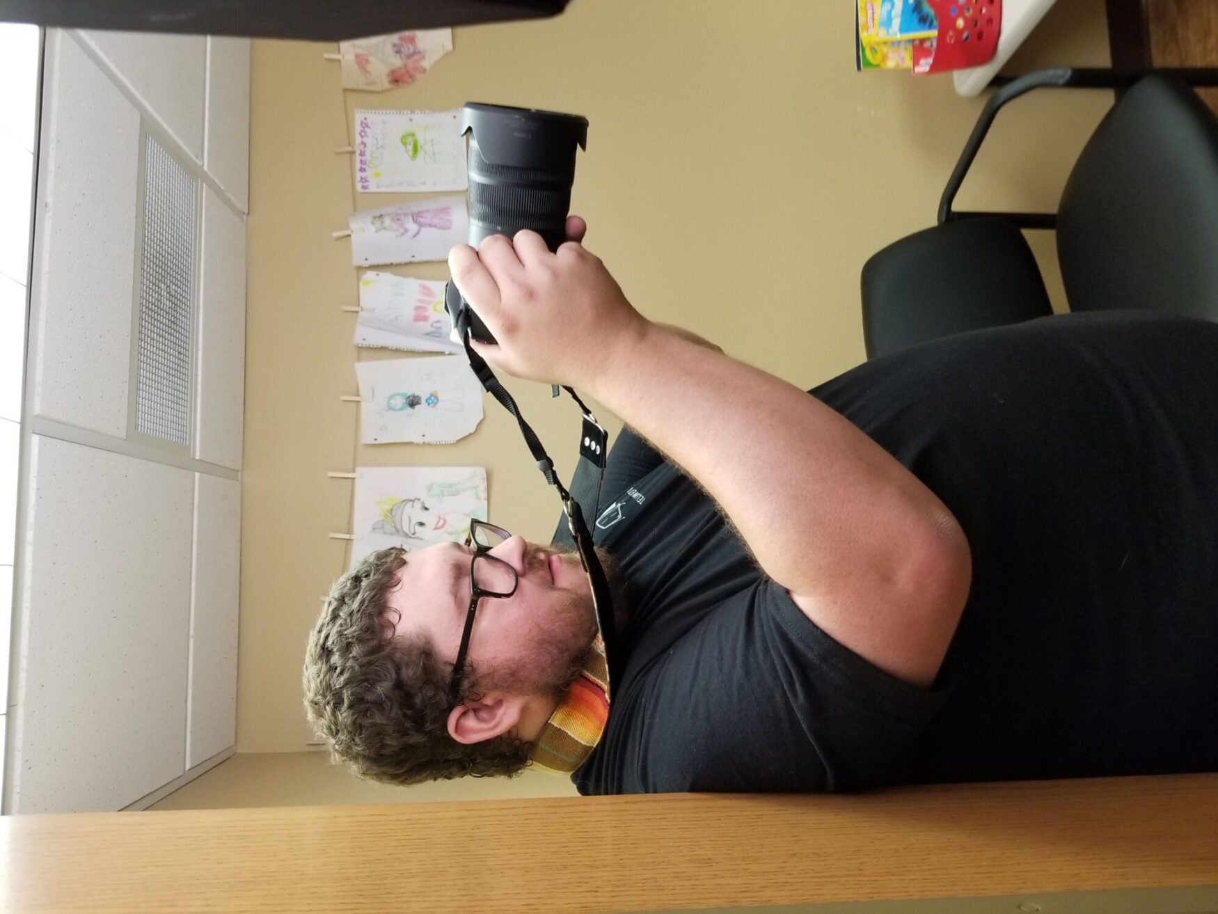 A man with glasses and a beard takes a photo in a room with children's drawings hanging on the wall. He is wearing a black shirt and a colorful camera lanyard.