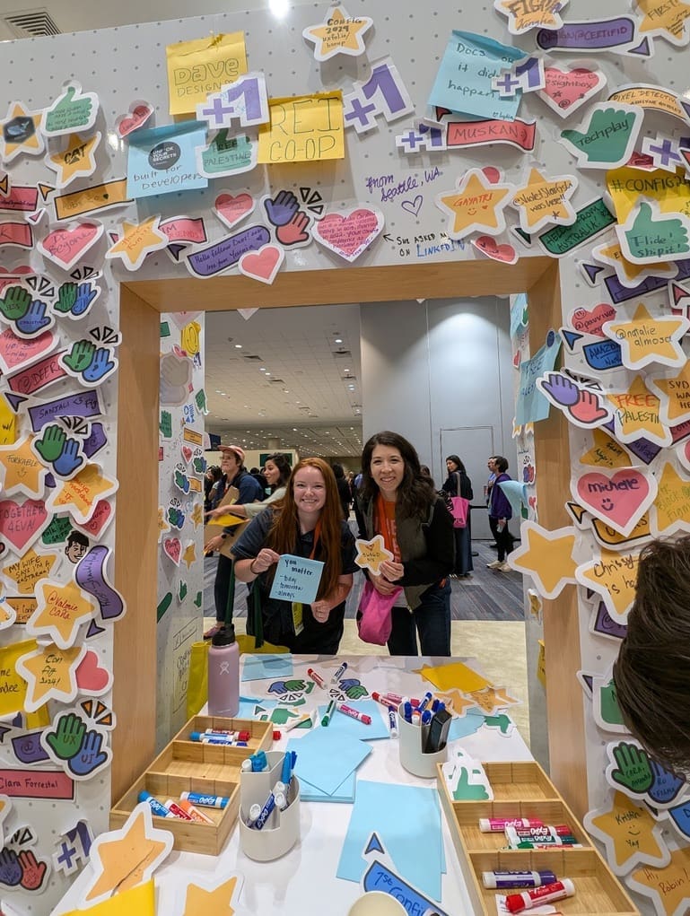 Two women standing at a colorful booth filled with sticky notes, stars, and hearts at a conference. They are holding a blue card that reads 'You matter,' smiling for the camera. The booth is covered with various handwritten messages and drawings, creating a vibrant and interactive display.