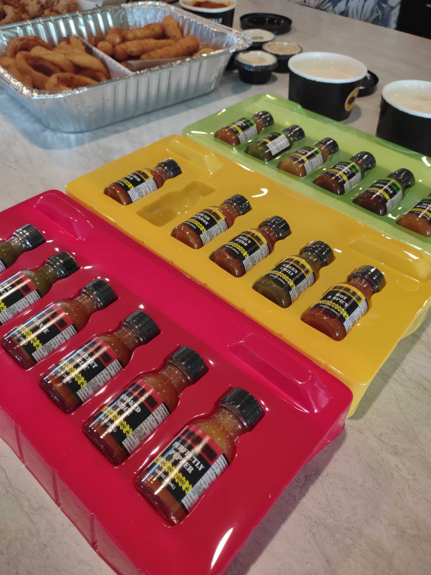 A close-up of a table with trays of assorted hot sauce bottles in vibrant red, yellow, and green packaging. In the background, there is a foil tray with fried appetizers and several small cups of dipping sauces, along with two cups of what appears to be beer or soda. The scene suggests a setup for a tasting event or casual gathering.
