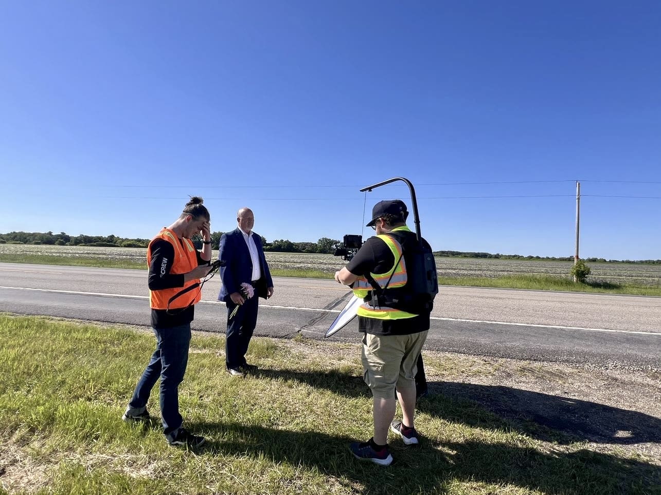 A film crew is working on the side of a road on a clear, sunny day. One crew member in an orange safety vest is looking down at equipment, another crew member in a yellow safety vest is operating a camera, and a man in a blue suit is standing in front of them holding flowers. The background features an open field with green grass and a clear blue sky.