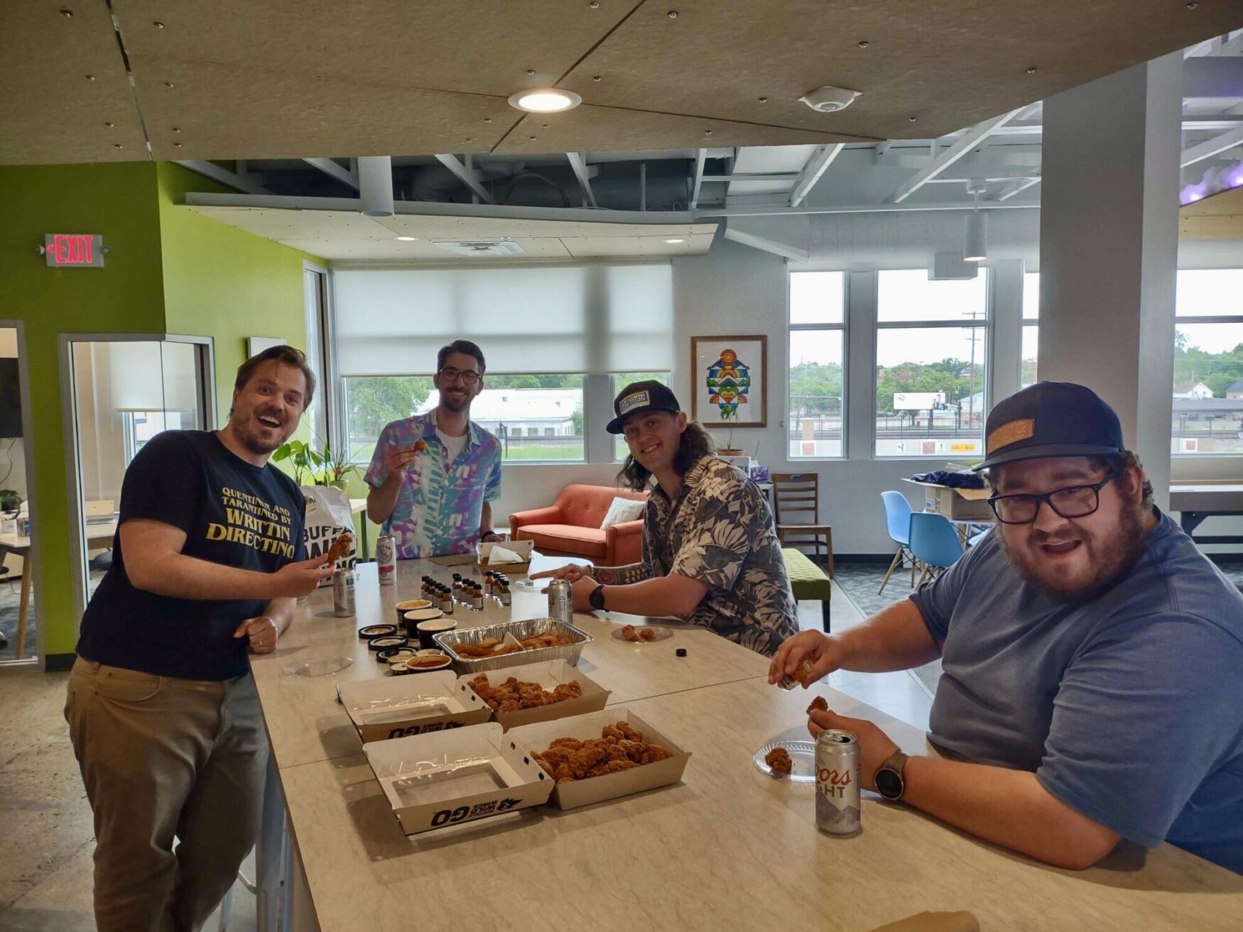 Four men standing around a kitchen island in a brightly lit office space, smiling and enjoying a spread of fried appetizers and various dipping sauces. They appear to be having a fun and casual gathering, with one person holding a can of Coors Light. The background shows modern office furniture and large windows with a view of the outside.