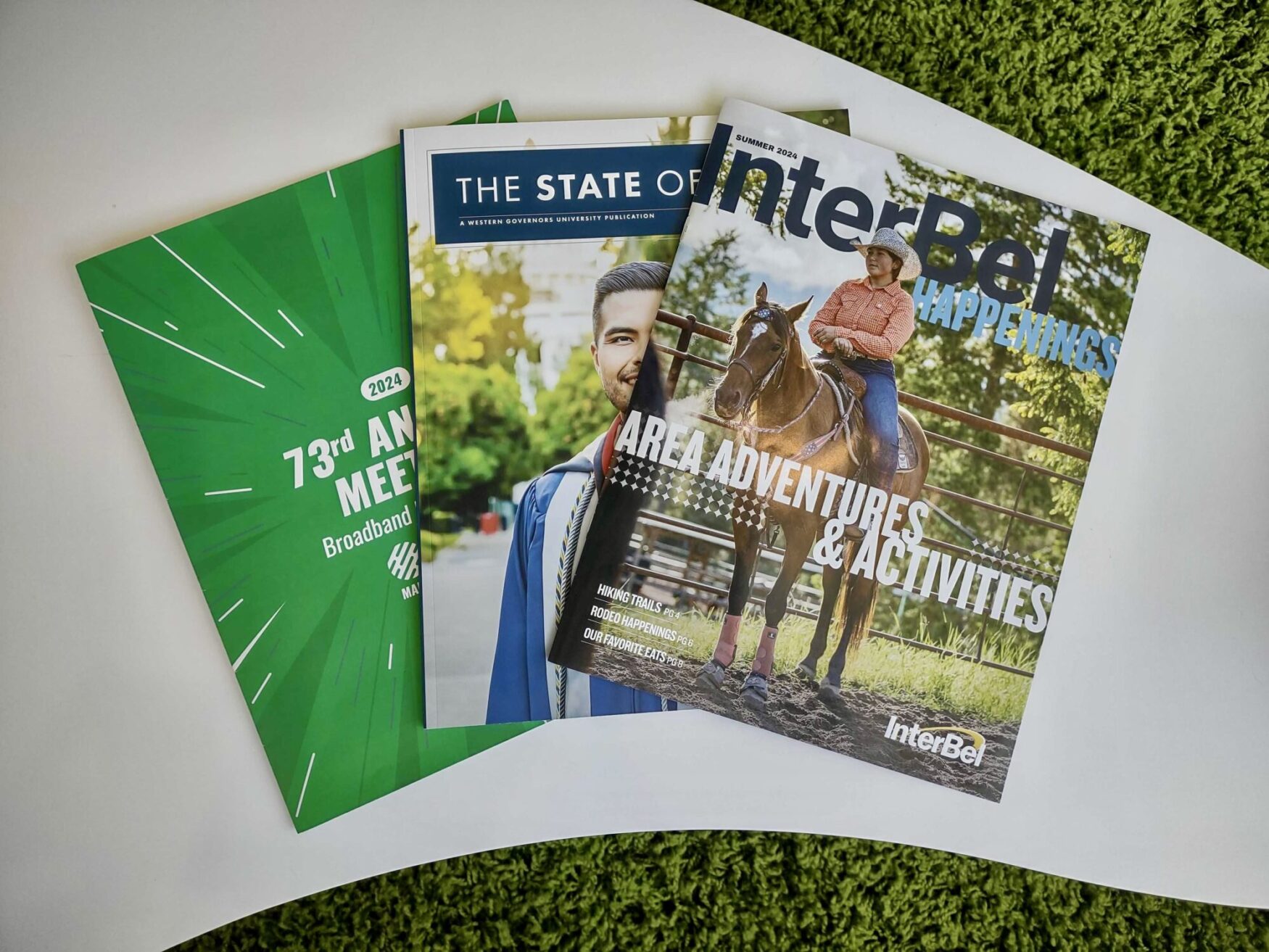 Three magazines are laid out on a white surface with a green carpet in the background. The leftmost magazine has a green cover with white text reading '73rd Annual Meeting 2024 Broadband' and other details. The middle magazine, titled 'The State of,' features a man in a graduation gown. The rightmost magazine, titled 'InterBel Happenings,' shows a person in a cowboy hat riding a horse with the subtitle 'Area Adventures & Activities.'