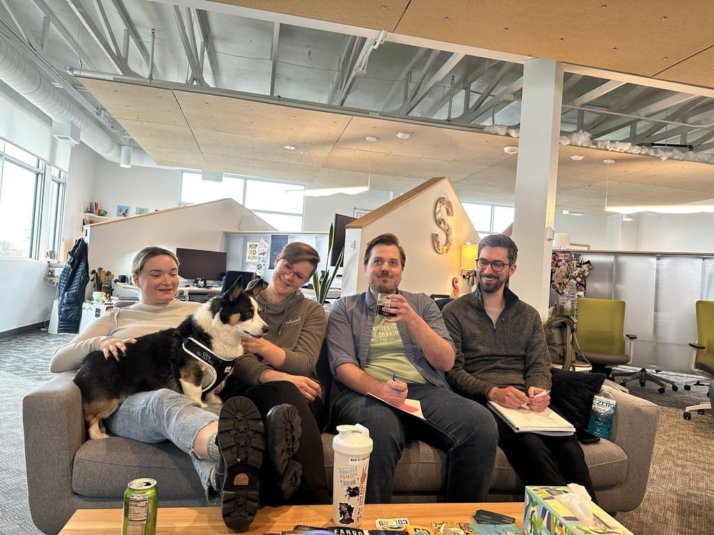 Four people sitting on a couch in a modern office space, smiling and relaxed. One person is holding a dog, adding a casual and friendly atmosphere. The background shows various office furniture and decor, creating a comfortable and collaborative environment.