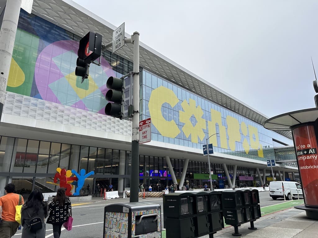 Exterior view of a large convention center with a colorful banner displaying 'CONFIG' in bold yellow letters. The building features glass walls and geometric designs, with people walking on the sidewalk in front. A traffic light and street signs are visible, along with public art sculptures near the entrance.