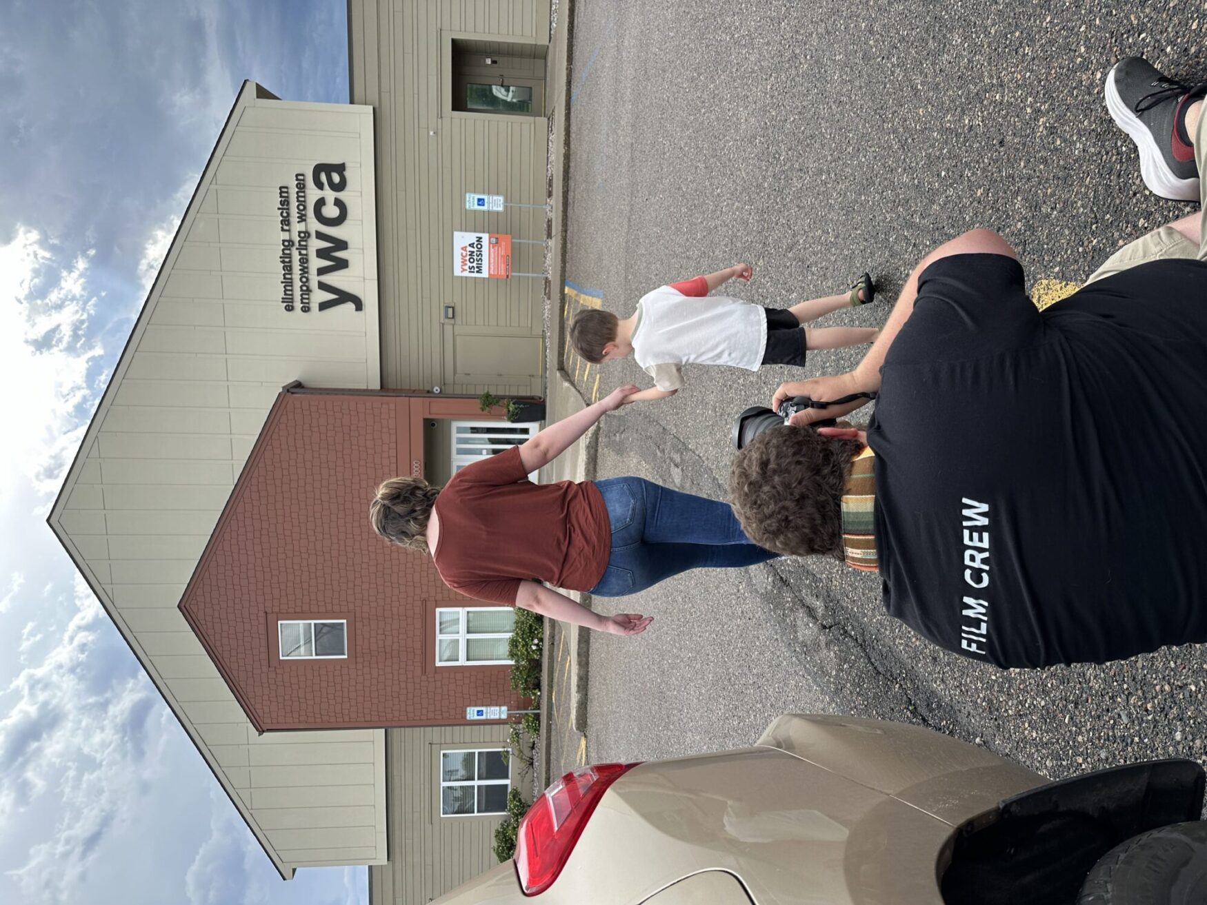 A film crew member in a black 'FILM CREW' shirt crouches to take a photo of a woman and child walking toward a YWCA building.