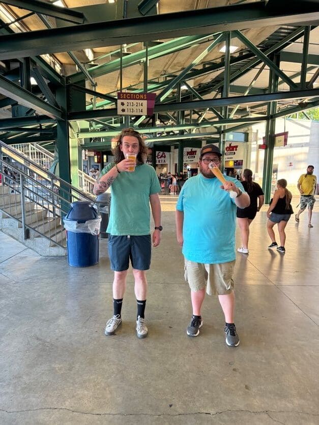 Two men in casual attire enjoy refreshments at a stadium. One is drinking from a cup, and the other is drinking from a comically large glass.