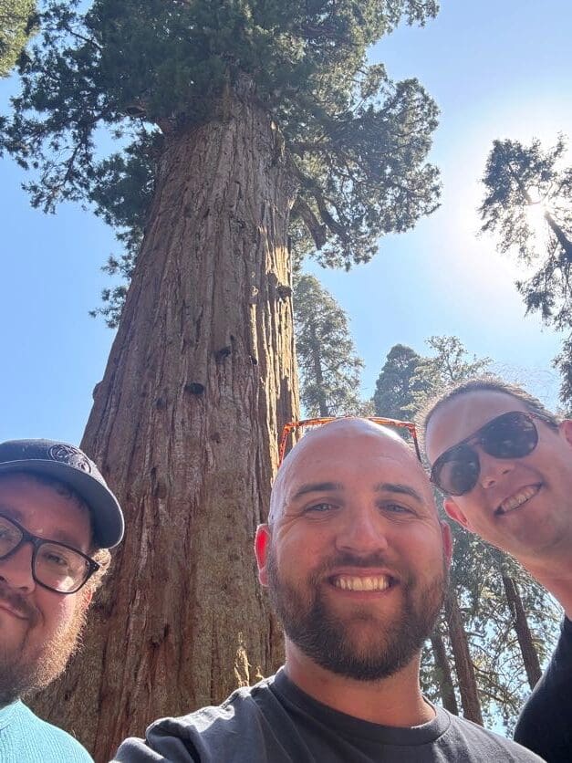 Three men take a selfie with a giant sequoia tree towering above them in the background. The sky is clear and sunny."