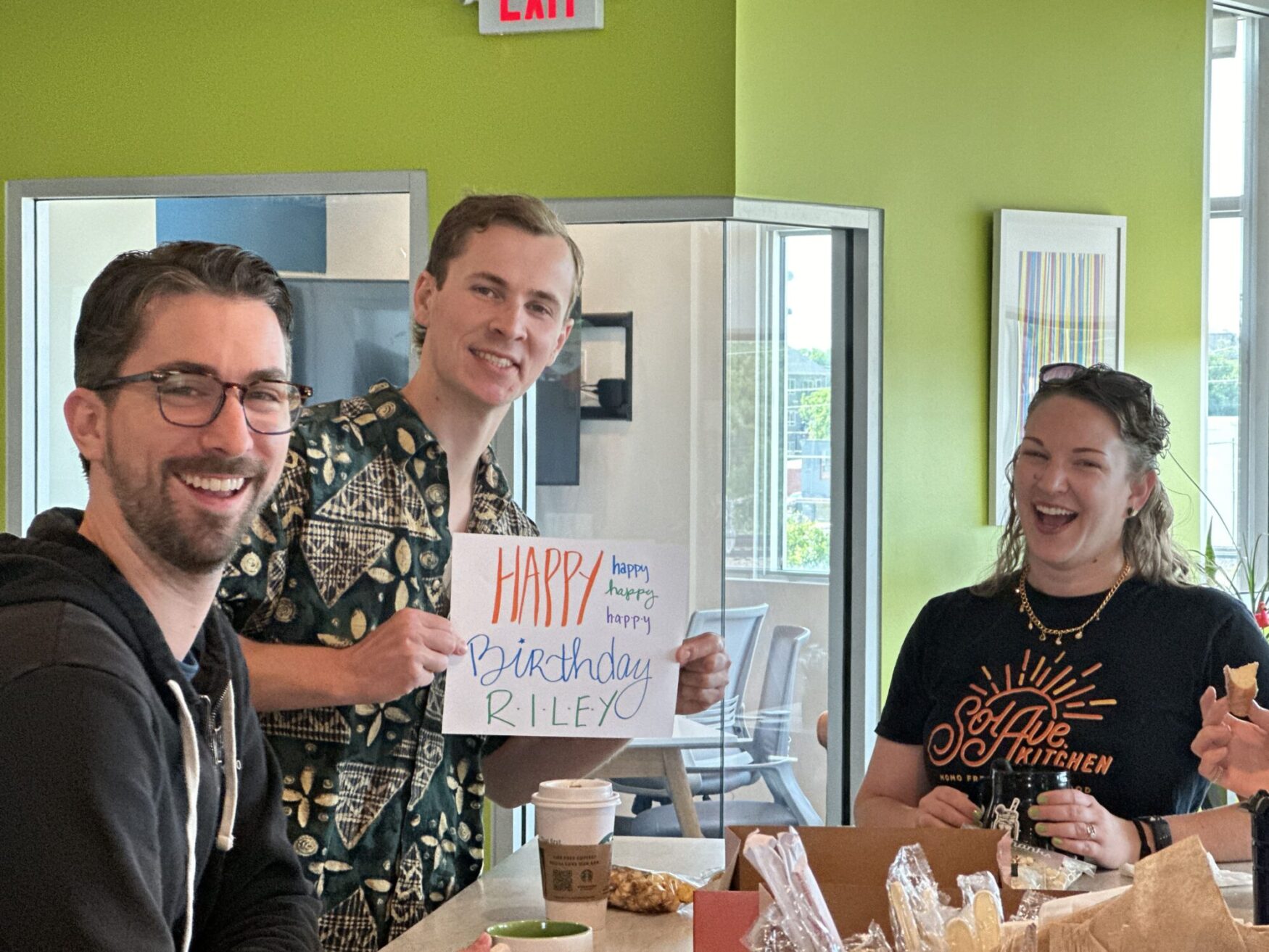 Three people in an office kitchen are smiling and celebrating. One person is holding a sign that says 'Happy Birthday Riley.' There are coffee cups and pastries on the table.
