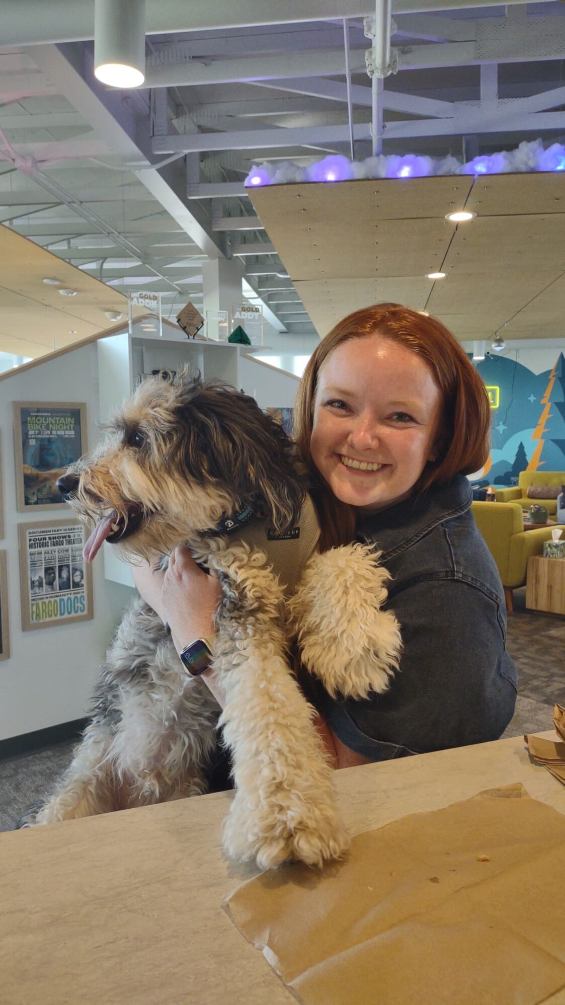 A woman with red hair smiles while holding a fluffy gray and white dog in an office with colorful decor and posters on the wall.